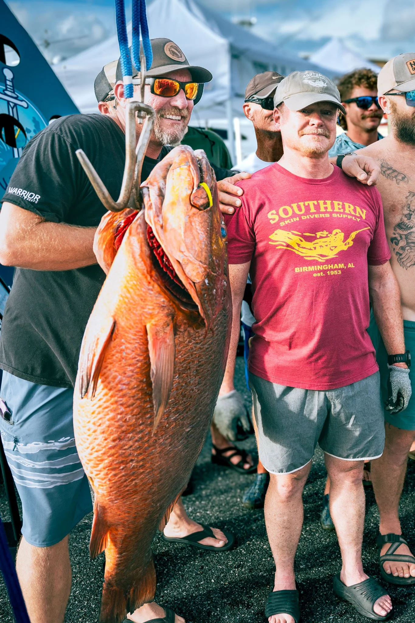Group of men at a fishing event, one holding a large cubera snapper with orange and gold scales, others standing nearby, some wearing sunglasses and hats, with tents in the background.