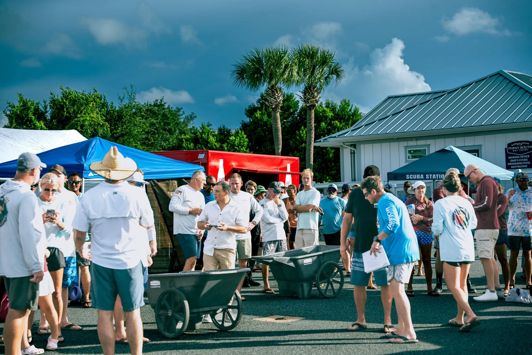 A group of people gathered at the Warrior Memorial Reefs Reels and Spears Fishing Tournament event at Point South Marina near tents and a building with palm trees in the background, some looking at their phones and others talking, on a sunny day.