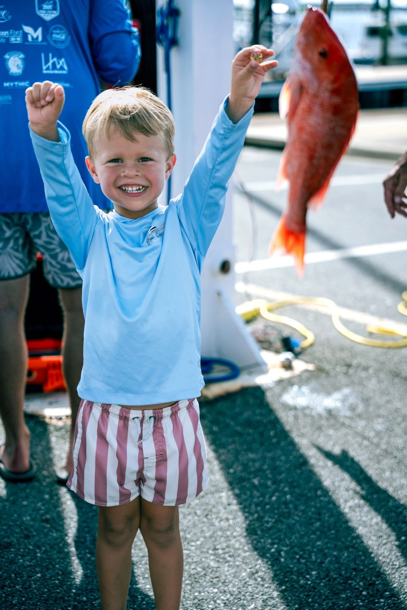 A young boy with short blonde hair, wearing a light blue long-sleeve shirt and pink and white striped shorts, smiling and raising his fists in excitement, standing outdoors near a red fish hanging.