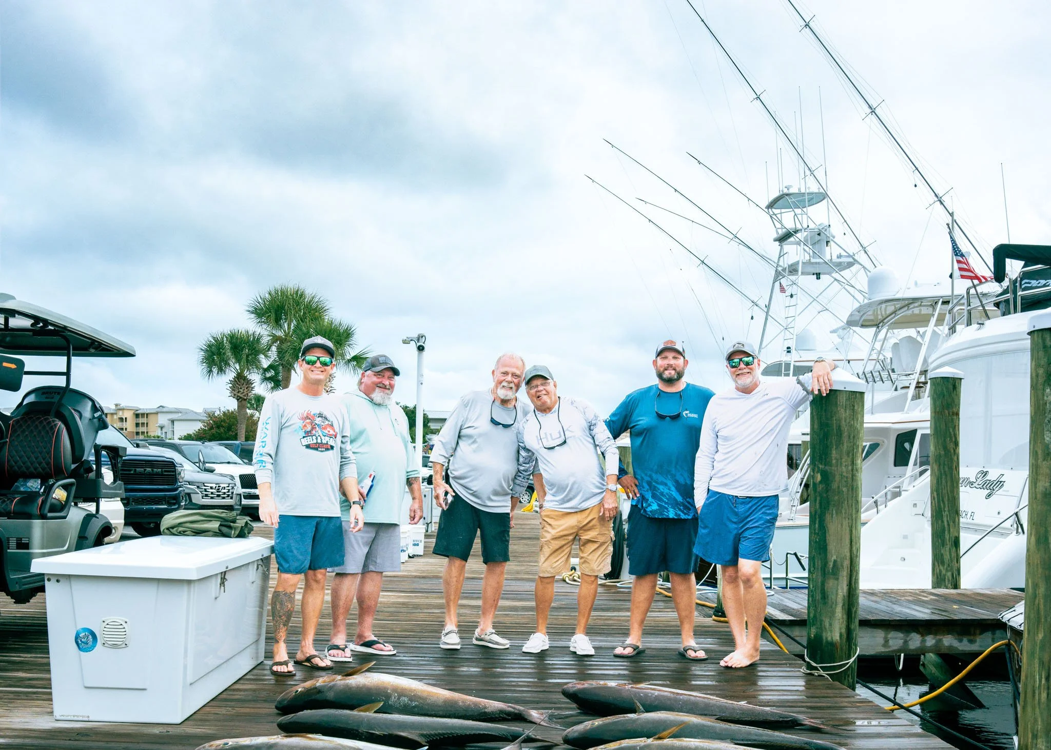 Group of six men standing on a dock at a marina, with boats and yachts in the background, and a large fish lying on the dock in front of them.