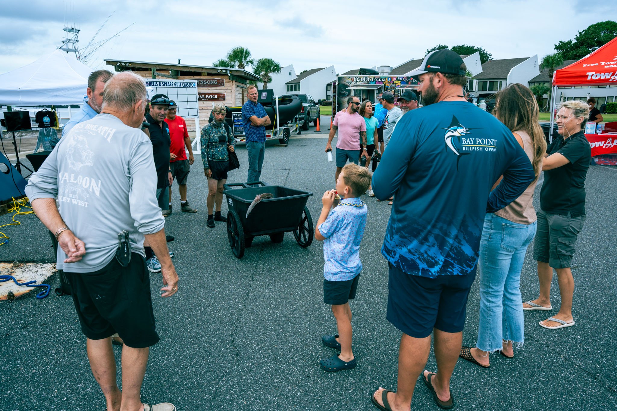 People gathered outdoors at a fishing event at Point South Marina - Bay Point (Panama City Beach), standing and talking near a tent and a whiteboard, with boats and houses in the background.