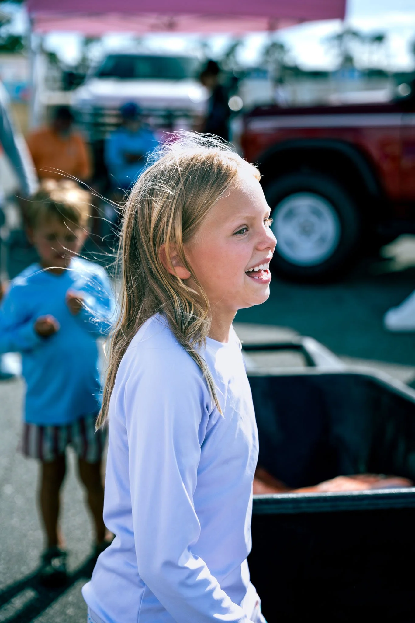 A smiling girl with blonde hair wearing a white shirt at an outdoor market, with people and vehicles blurred in the background.