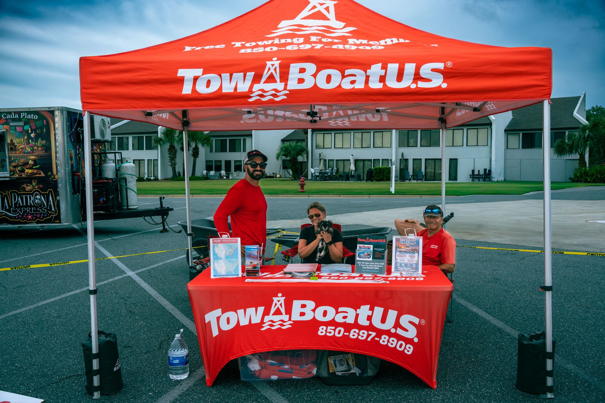 A promotional booth for TowBoatU.S. with a red canopy, two men and a woman sitting at a table, and a dog. The booth has brochures and signs, with a building and palm trees in the background.