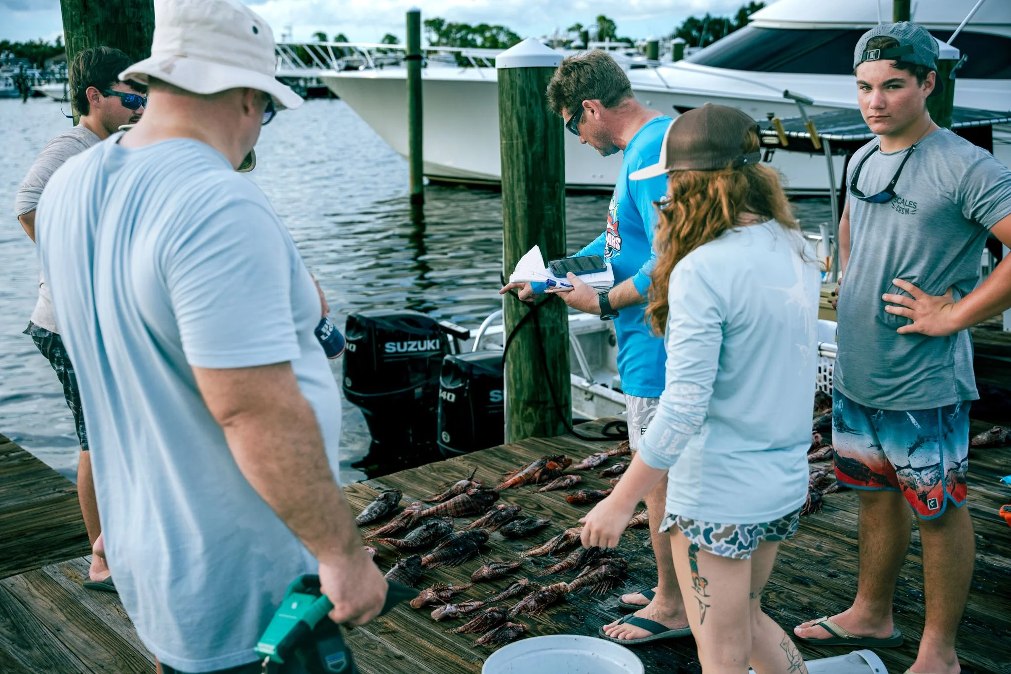 Group of young people and adults standing on a wooden dock with lobsters laid out on the surface, near boats docked in a marina, some holding clipboards and wearing casual summer clothing and hats.