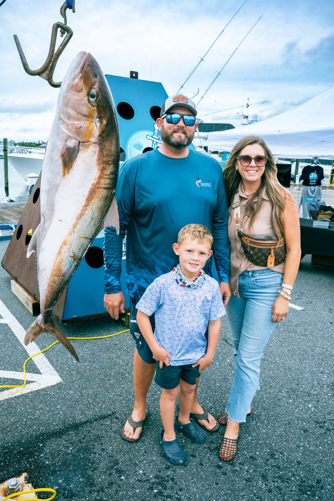 A man, woman, and two children standing on a parking lot in front of a large fish mounted on a wooden display. The man wears sunglasses and a blue shirt, the woman wears sunglasses and a beige top, one child wears a floral shirt and shorts, and the o