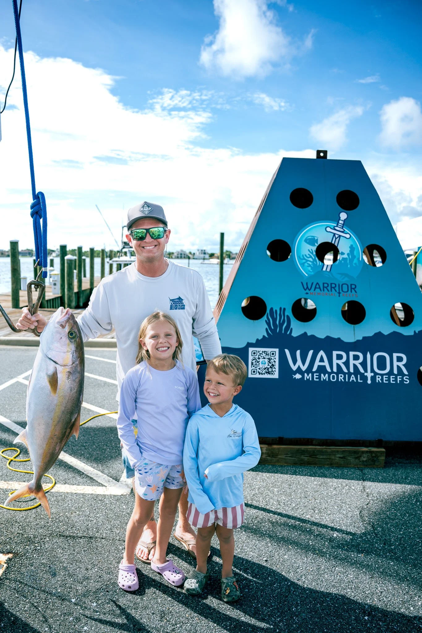 A smiling man holding a large fish with two children standing in front of a sign for Warrior Memorial Reefs at Point South Marina - Bay Point (Panama City Beach). The background shows a dock and boats under a partly cloudy sky.