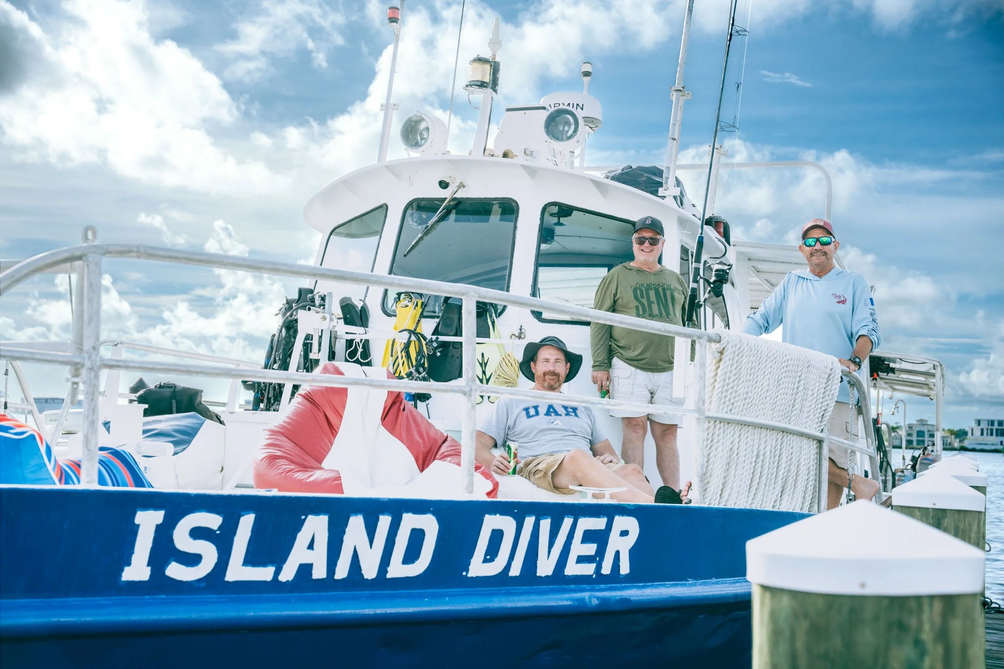 Four men on a boat with a sign reading "ISLAND DIVER"; the boat has diving equipment and the men are wearing sunglasses and casual shirts, with a background of a partly cloudy sky.
