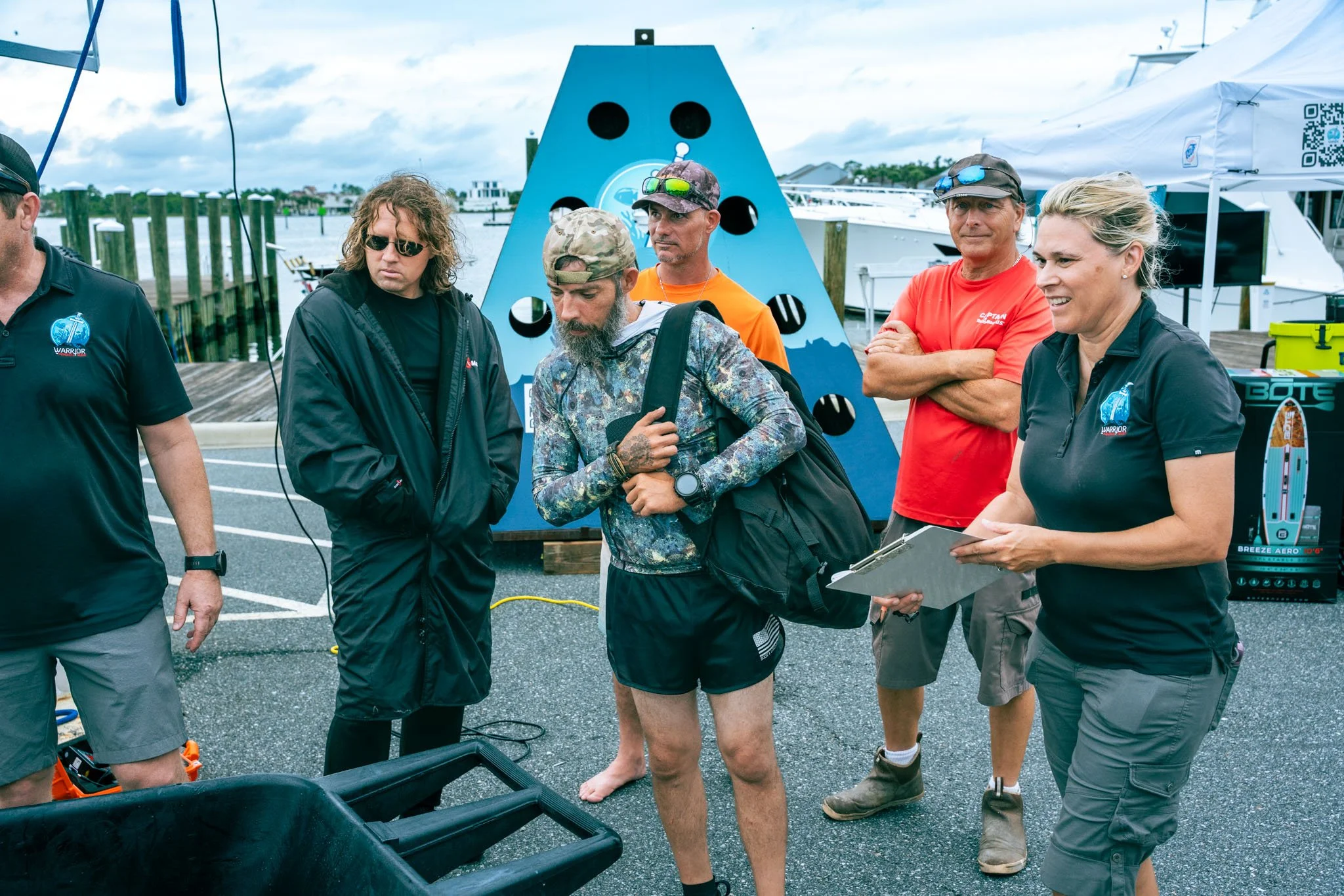 Group of people gather outdoors near a dock, engaging in a discussion or briefing. Several individuals are dressed casually, some with sunglasses and hats, and one person holds a clipboard. In the background, boats are docked, and a blue climbing wal