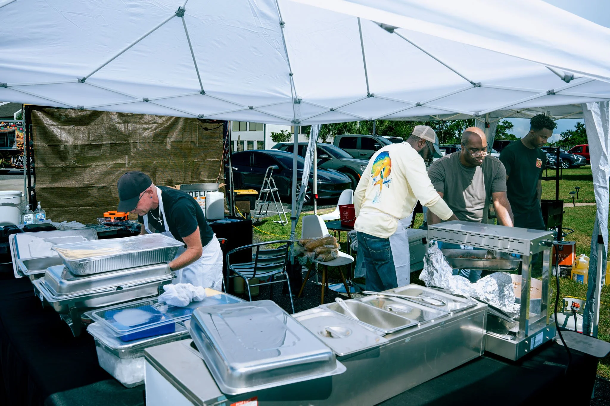 People preparing food at an outdoor event at Point South Marina - Bay Point (Panama City Beach) under a white canopy tent, with porches, parked cars, and greenery in the background.
