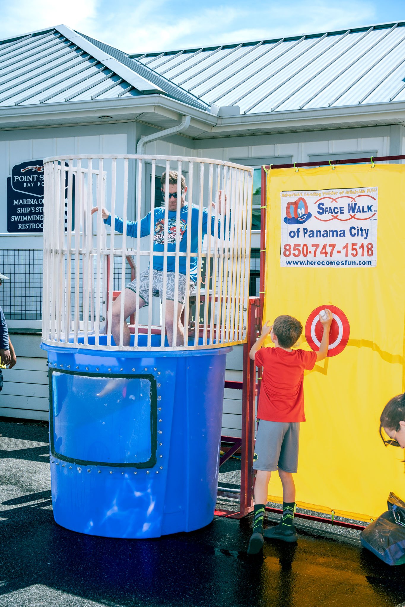 A young boy wearing a red shirt and gray shorts playing a carnival game at Space Walk of Panama City, with another person on a dunk tank behind him and a yellow backdrop with a target.