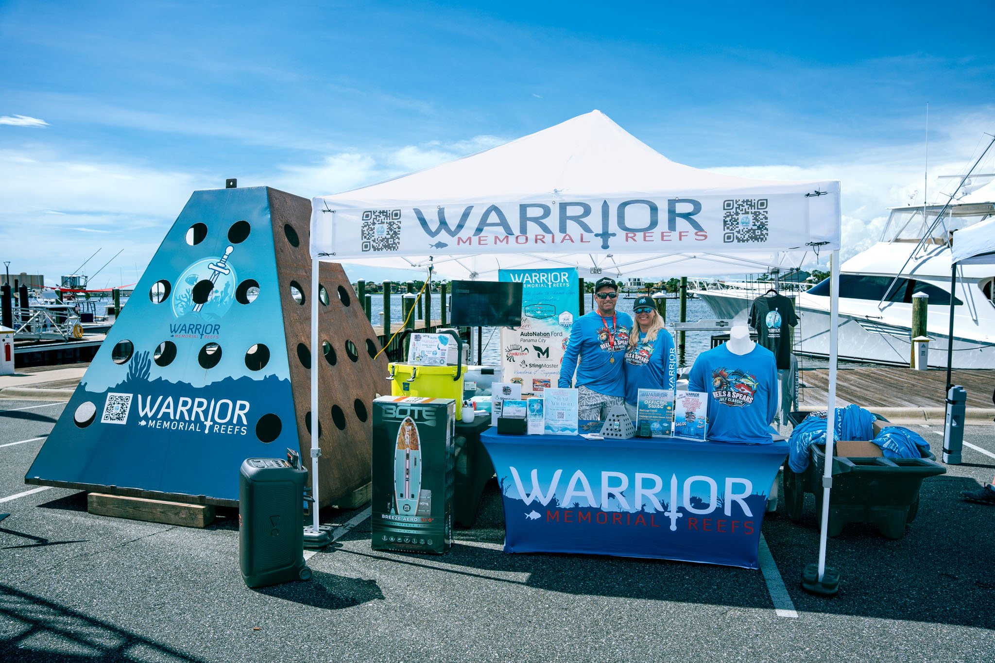 Booth at a marina promoting Warrior Memorial Reefs with banners, apparel, and informational materials, featuring two people smiling and standing behind the table.
