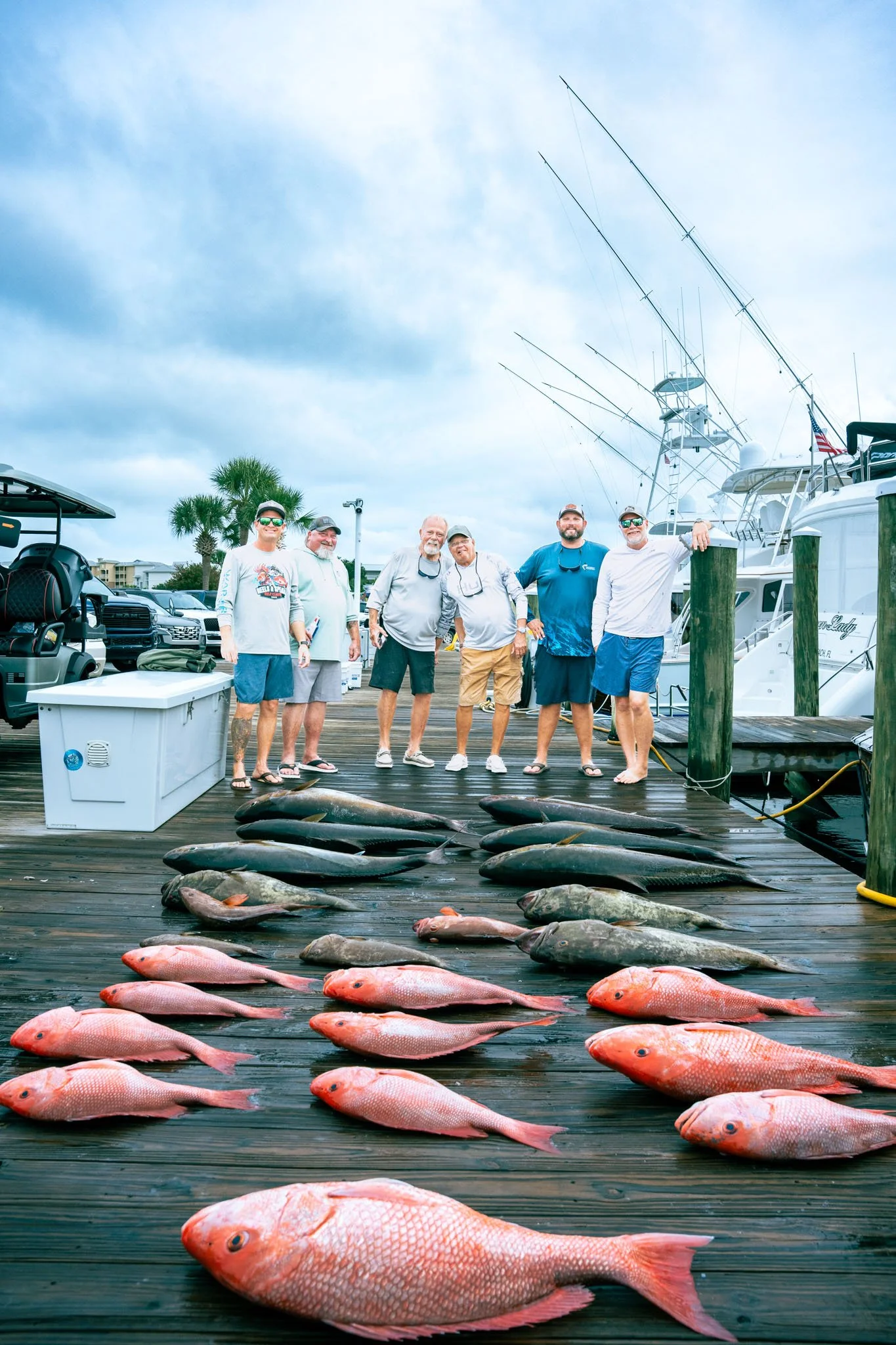 Group of six men standing on a dock with caught fish lined up in front of them and boats in the background.