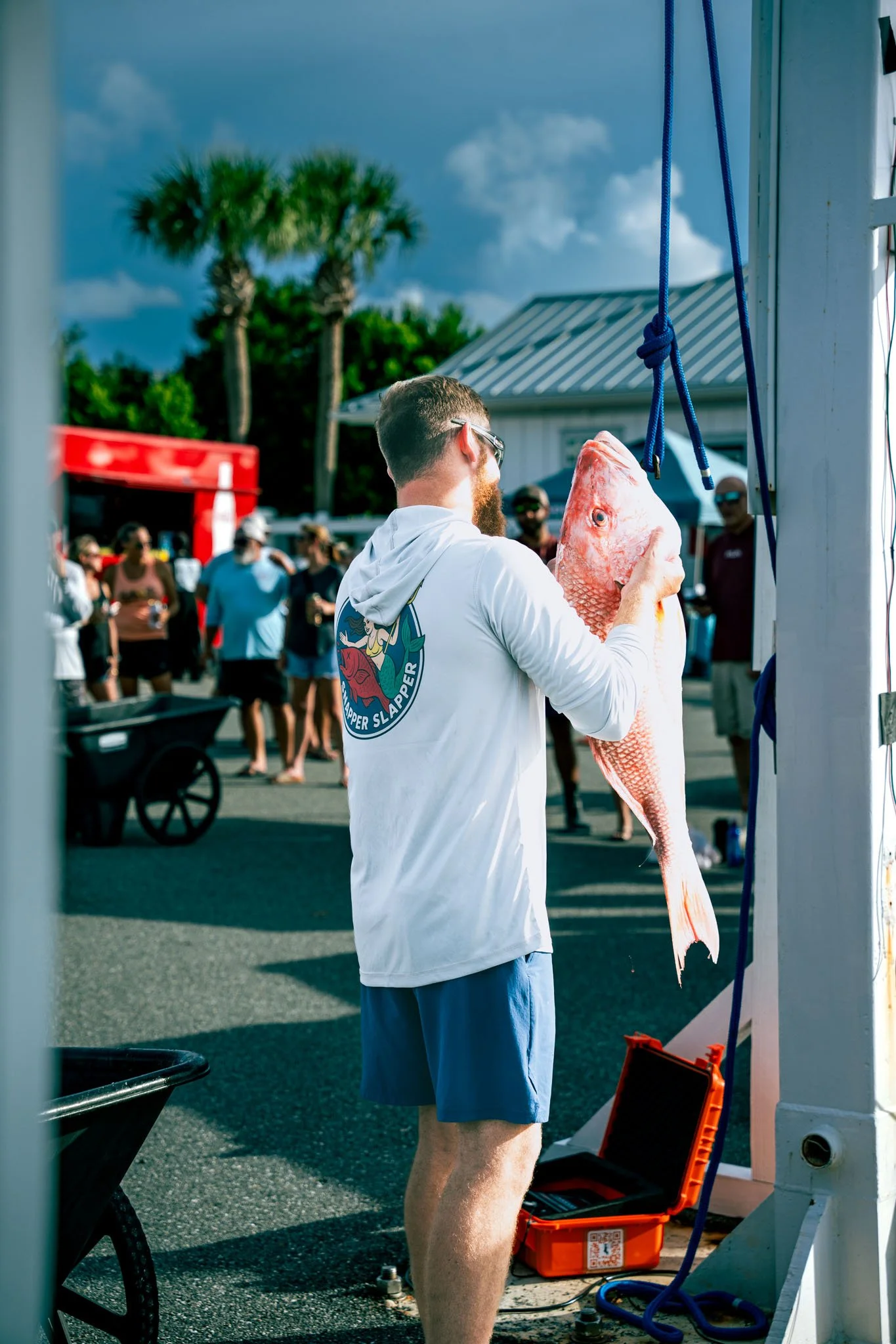 A man with sunglasses and a beard holding a large fish in front of a group of people at the Warrior Memorial Reefs Reels and Spears Fishing Tournament event at Point South Marina - Bay Point (Panama City Beach) on a sunny day.