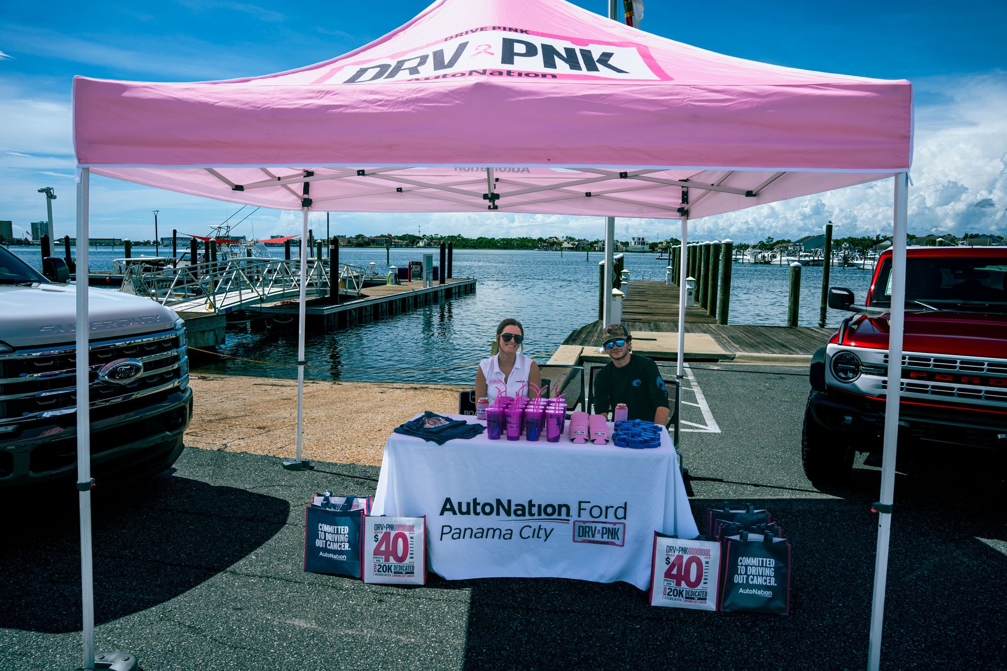A pink canopy with 'DRV PNK' logo and related text, set up at a waterfront parking area with boats and buildings in the background. Under the canopy, a table displays pink tumblers, wristbands, and flyers for AutoNation Panama City. Two smiling peopl