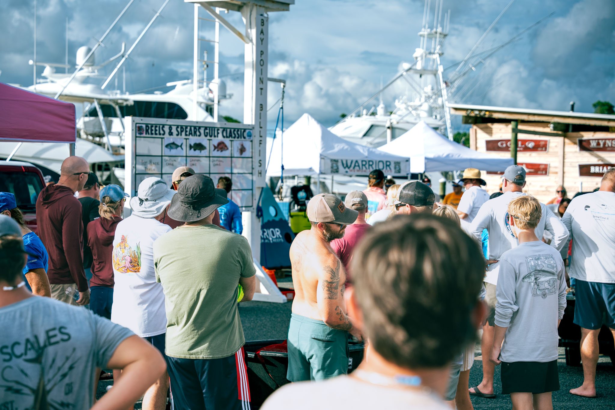 Crowd gathered at the Warrior Memorial Reefs Reels and Spears Fishing Tournament event at Point South Marina - Bay Point (Panama City Beach) with yachts and tents in the background.