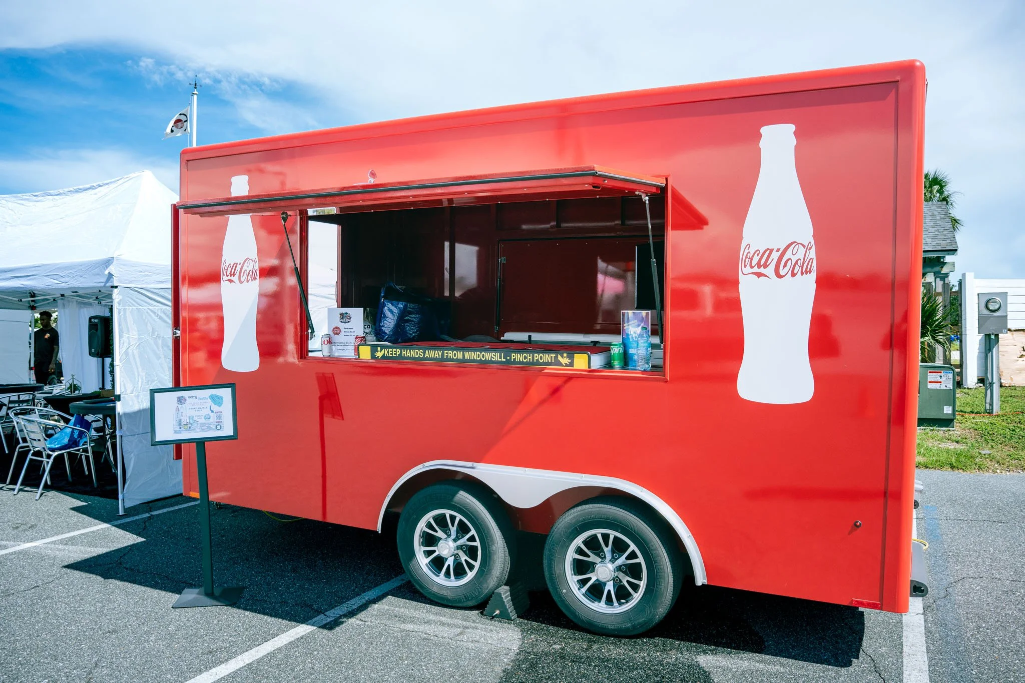 Red Coca-Cola branded food trailer with an open service window, located in an outdoor parking lot with tents and people nearby.