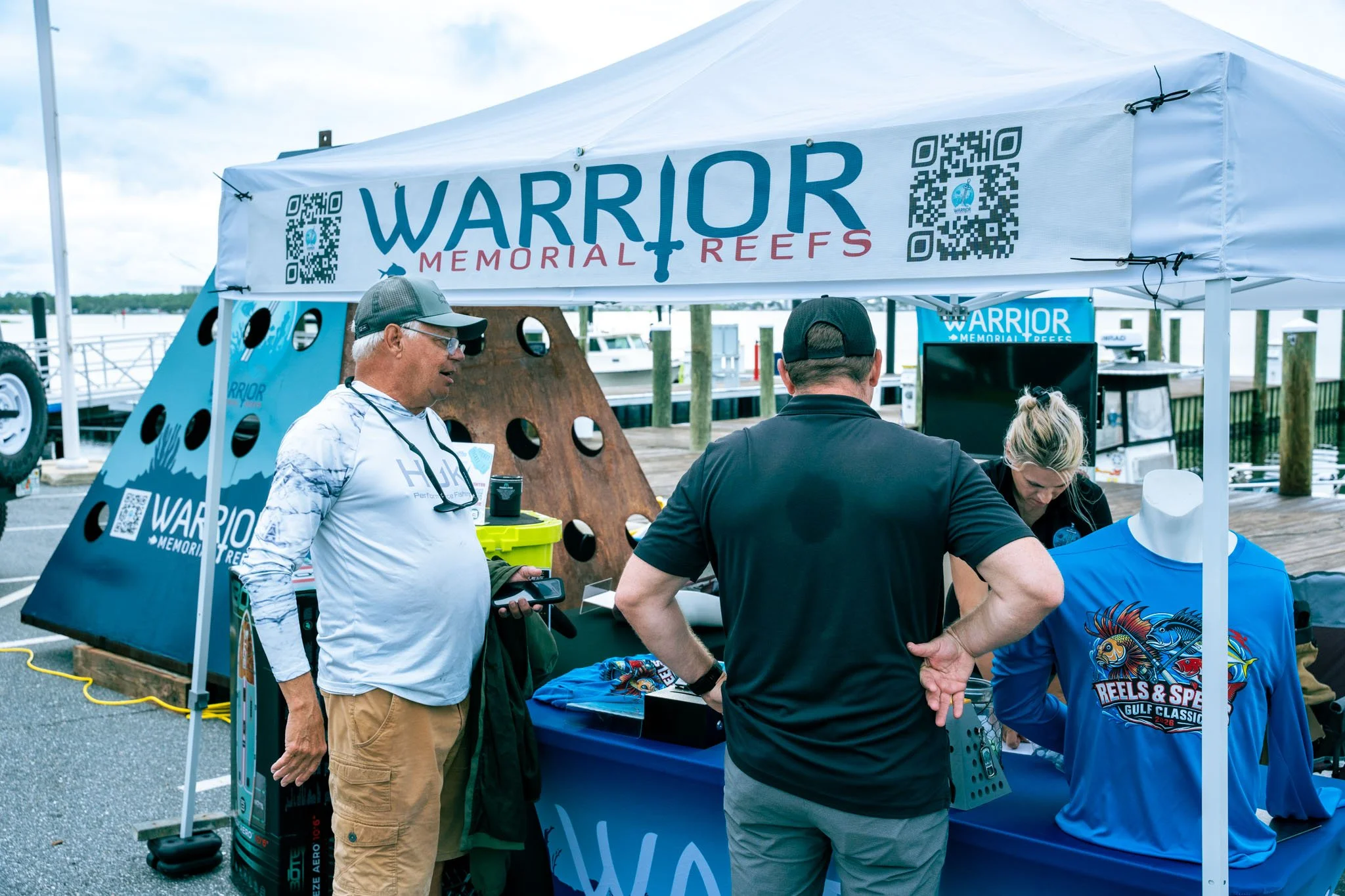 A group of people gathered under a white tent with a banner reading 'Warrior Memorial Reefs,' near a marina. The scene appears to be at an outdoor event.
