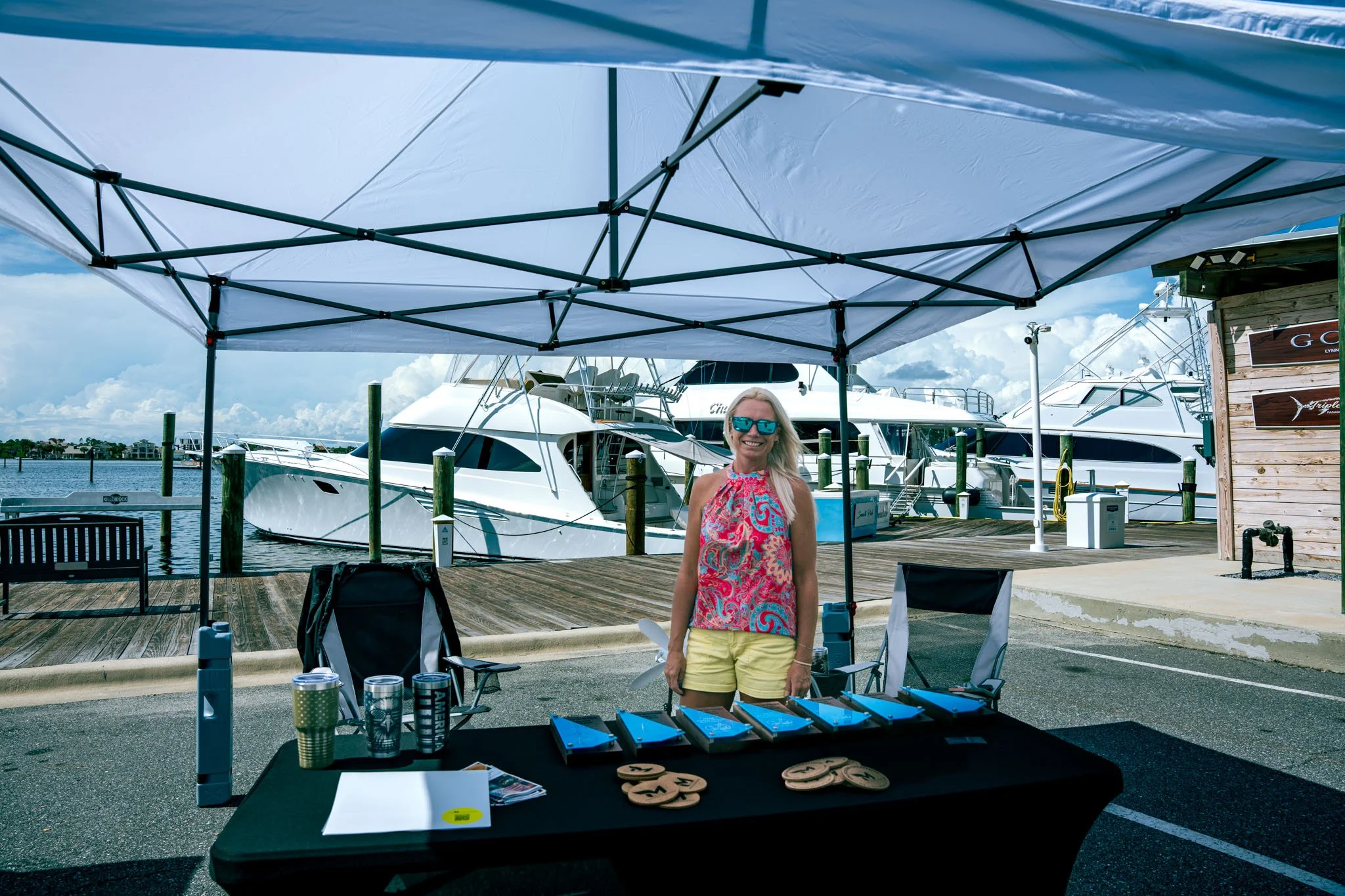 Woman standing at outdoor booth at Point South Marina - Bay Point (Panama City Beach) with sunglasses and colorful top, yachts in the background. 