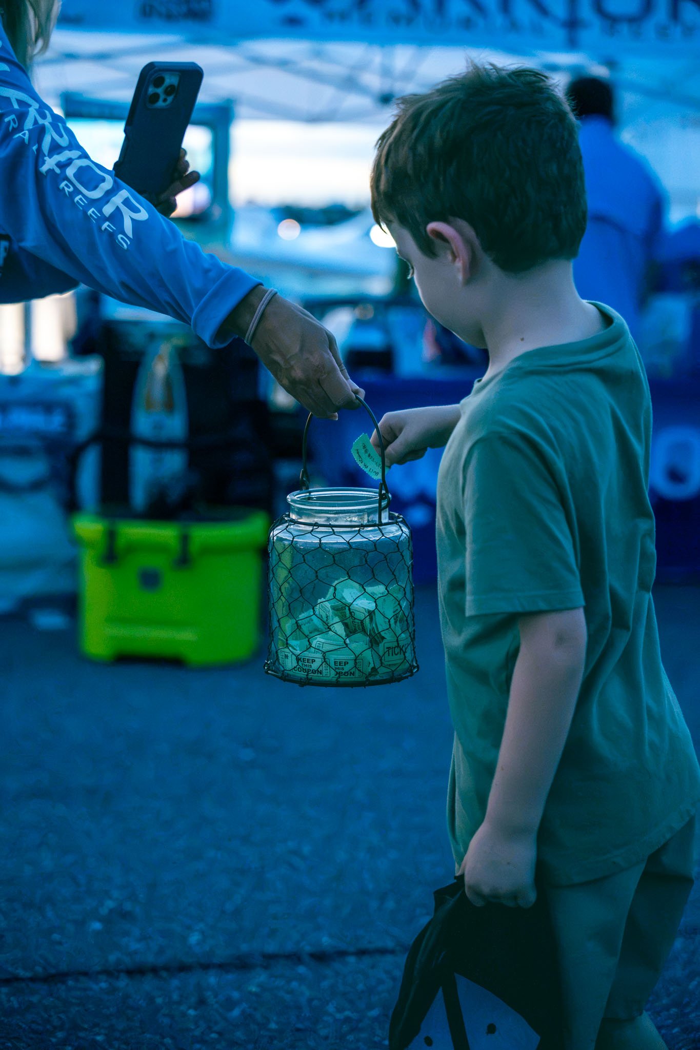 A young boy with brown hair and wearing a yellow t-shirt holds a wire lantern with a candle in it. An adult in blue clothing is handing him an object, possibly a ticket or note. The background has blue lighting and blurred objects, indicating an outd