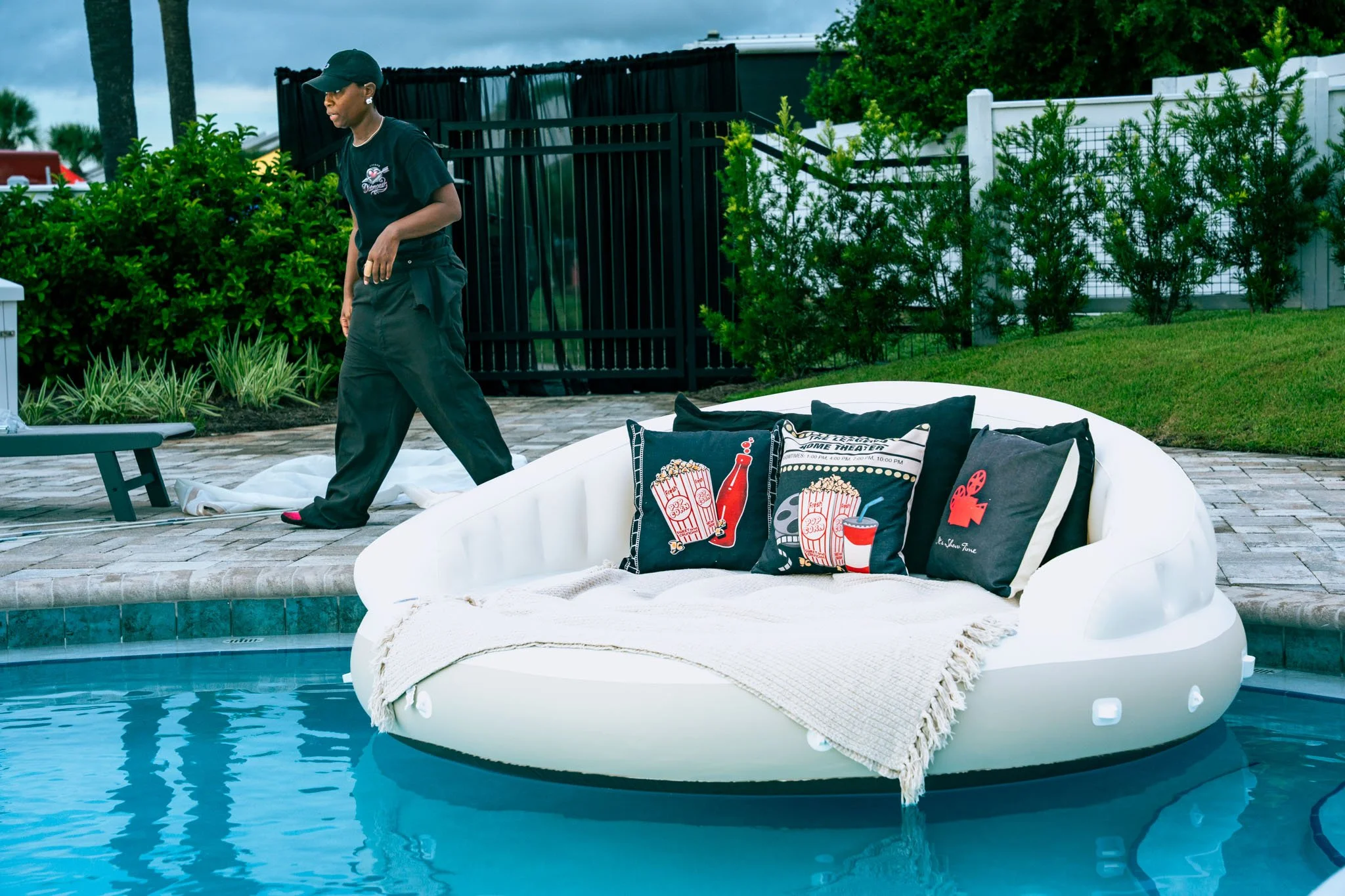 A person walking near a poolside with an inflatable raft decorated with images of popcorn, soda, and a film camera, surrounded by outdoor furniture and greenery.