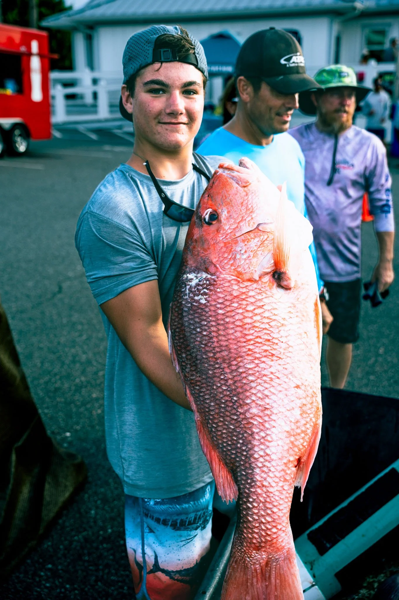 A young man wearing a blue cap and a gray shirt is holding a large red fish at a fishing location. Two men in hats stand behind him, and there are other people and vehicles in the background.