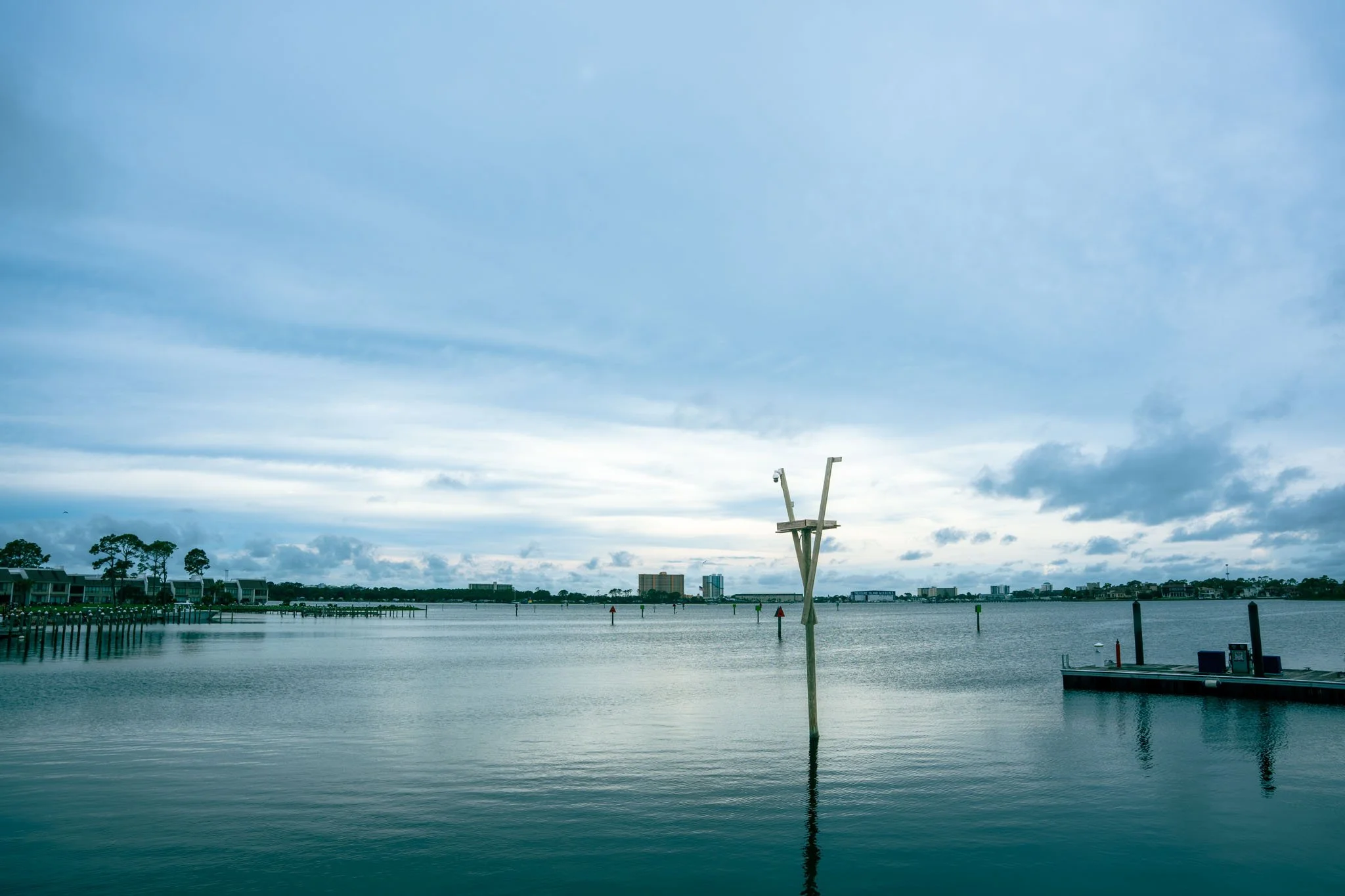 Grand Lagoon, Point South Marina - Bay Point (Panama City Beach) with a wooden channel marker in the water, a wooden pier, and distant buildings under a cloudy sky.