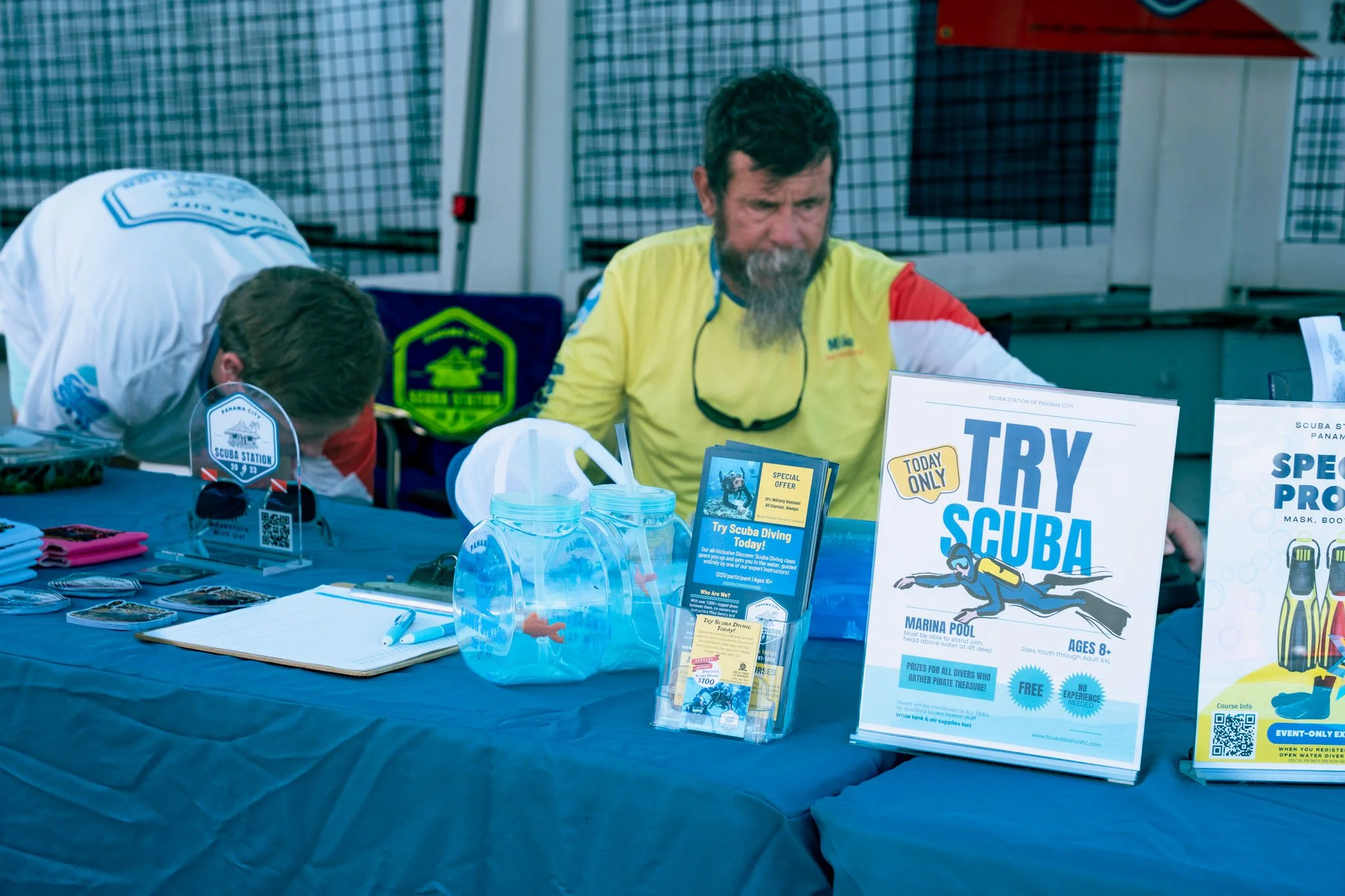 A table at a scuba diving event displays promotional materials, signs, and small items such as pamphlets, a water bottle, and a notebook. Behind the table, two men are seated, one with a beard and wearing a yellow shirt, focused on the materials. The