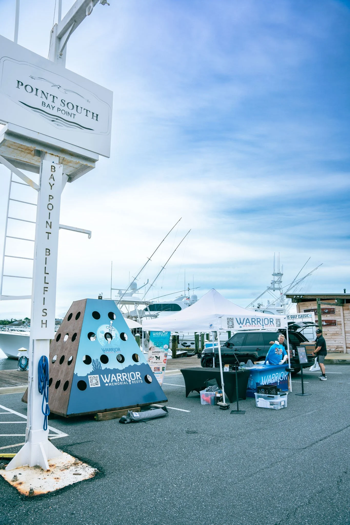 Point South Marina - Bay Point (Panama City Beach) with boats in the background and a booth set up with a blue banner that says Warrior Memorial Reefs. There are a few people at the booth and a tent providing shade.