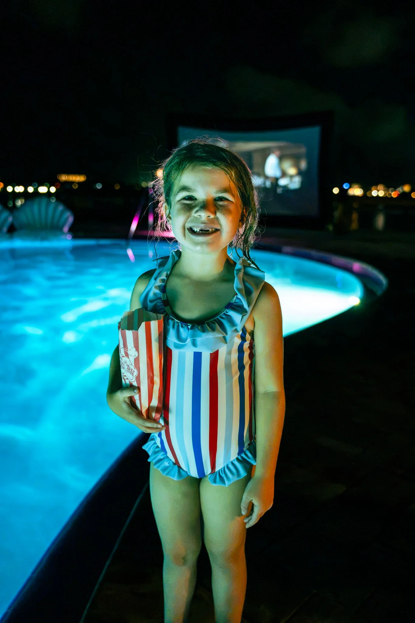 A young girl in a striped swimsuit smiling by a lit-up Point South Marina - Bay Point (Panama City Beach) pool at night, with a movie screen visible in the background.