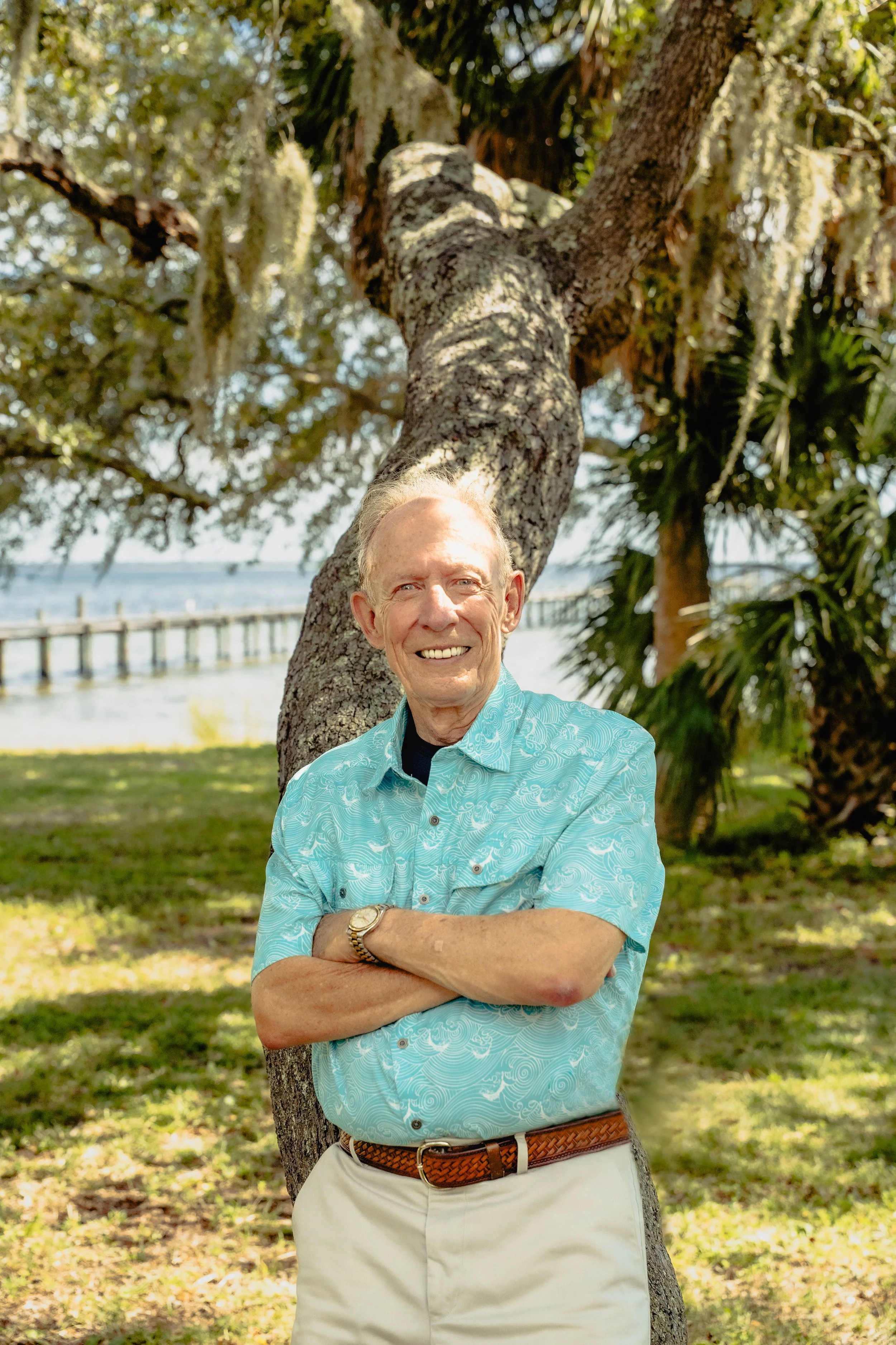 An older, well-dressed caucasian man in a turquoise patterned shirt and khaki pants, leaning against an oak tree in a park. The ocean is directly behind him with a long dock extending out on the left side of the image.