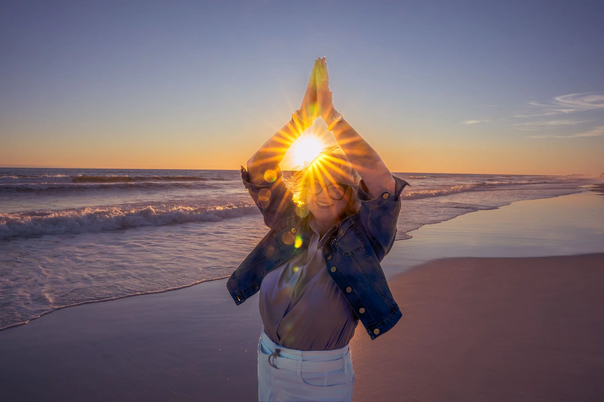 Woman practicing yoga or meditation on the beach during sunset, with her hands in prayer position and sun rays shining through her hands.