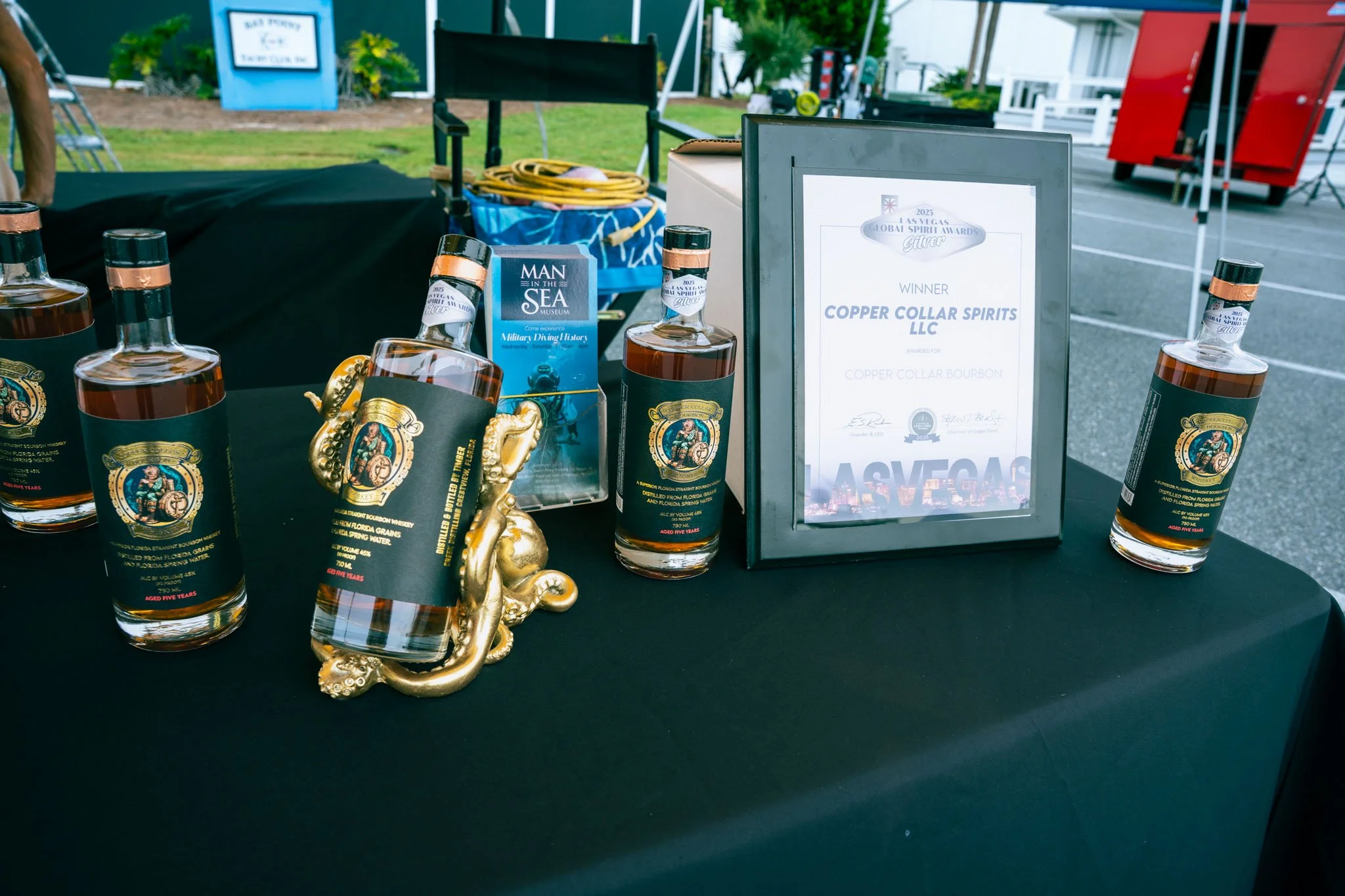 Display of whiskey bottles, a framed award certificate for Copper Collar Spirits LLC, and a small golden souvenir, on a black table at an outdoor event.
