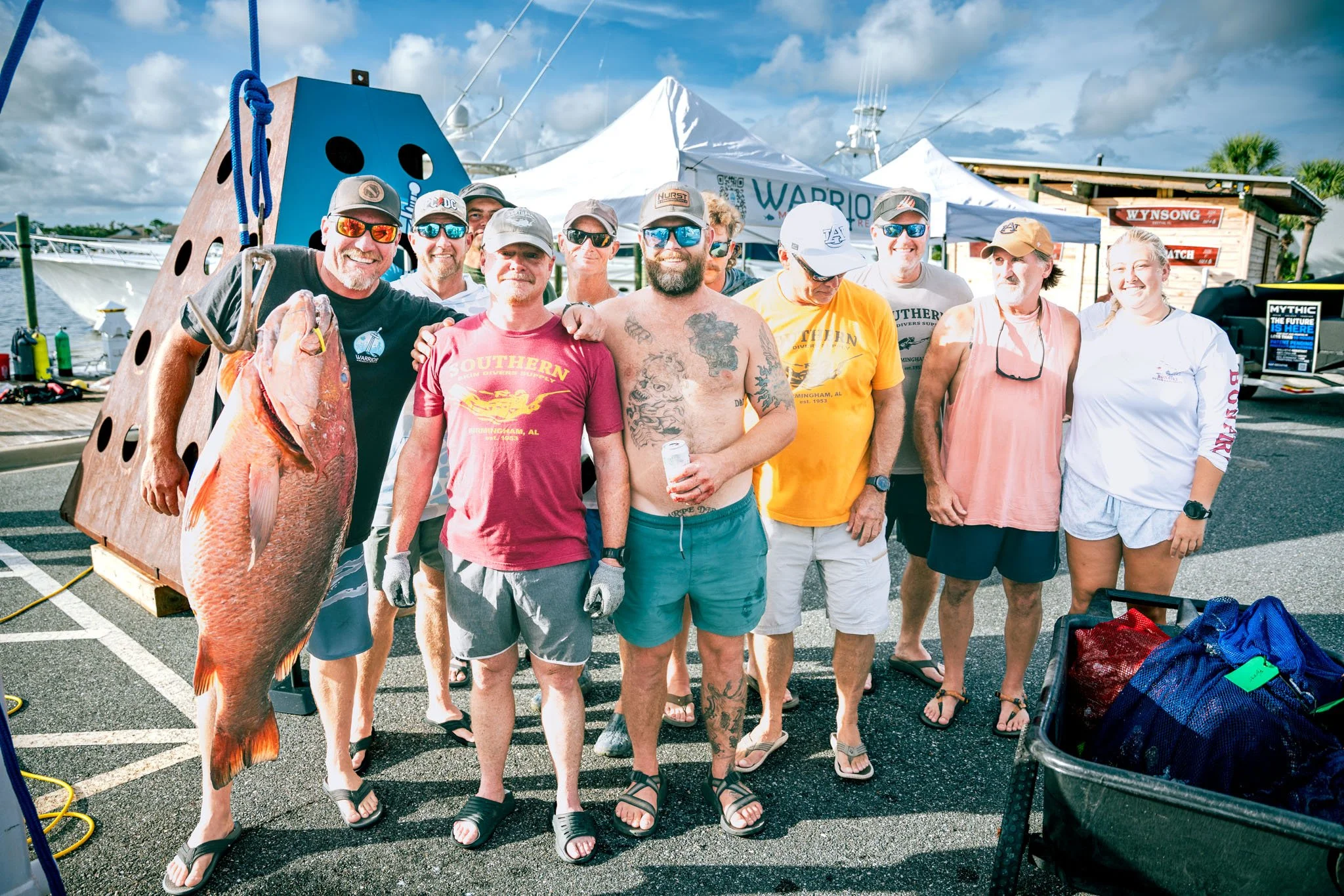 Group of people standing together outdoors, some wearing sunglasses, with a large cubera snapper hanging from a wooden platform, and boats and tents in the background on a partly cloudy day.