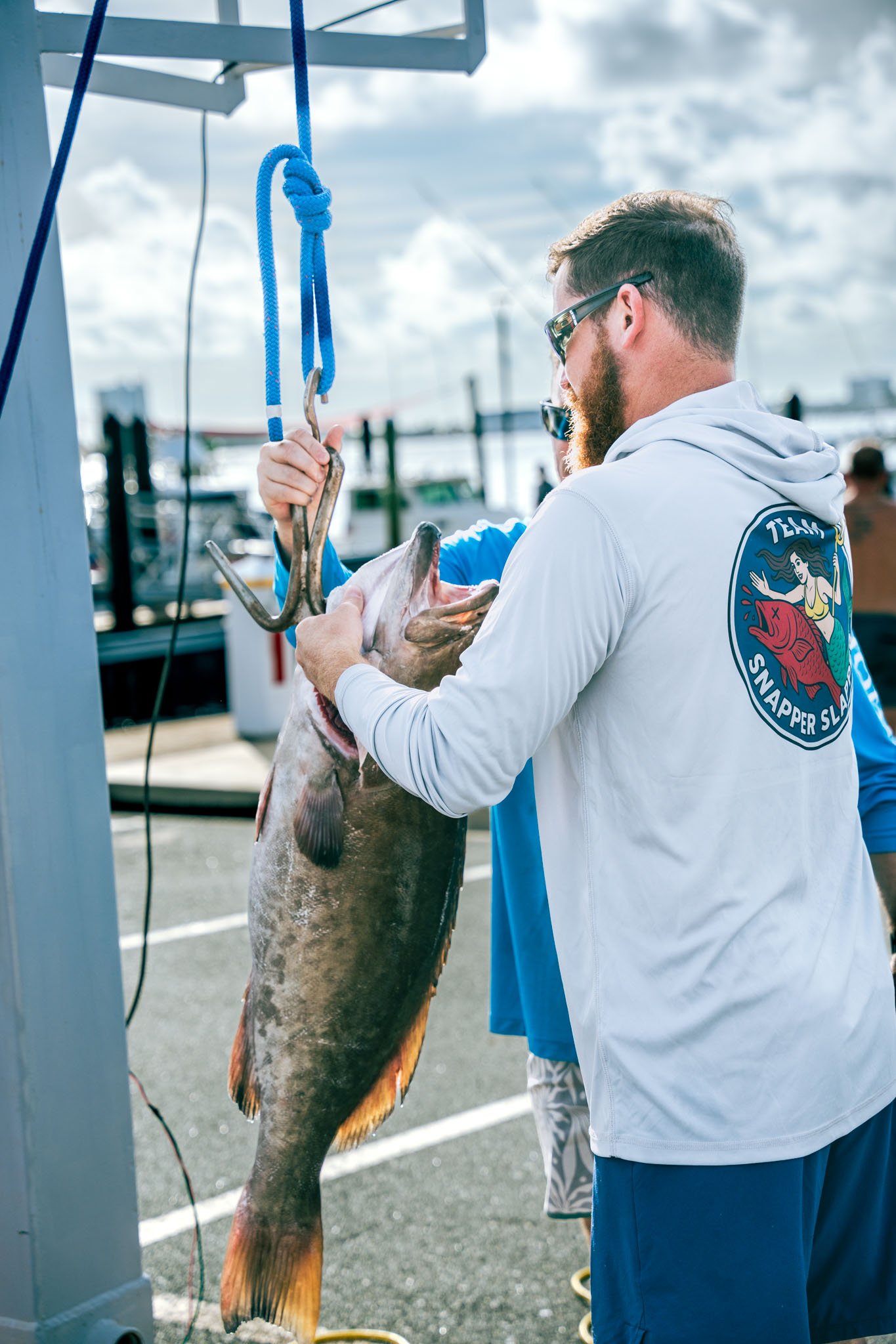 A man with sunglasses and a hoodie holding a large gag grouper suspended from a weighing scale at Point South Marina - Bay Point (Panama City Beach), with boats and cloudy sky in the background.