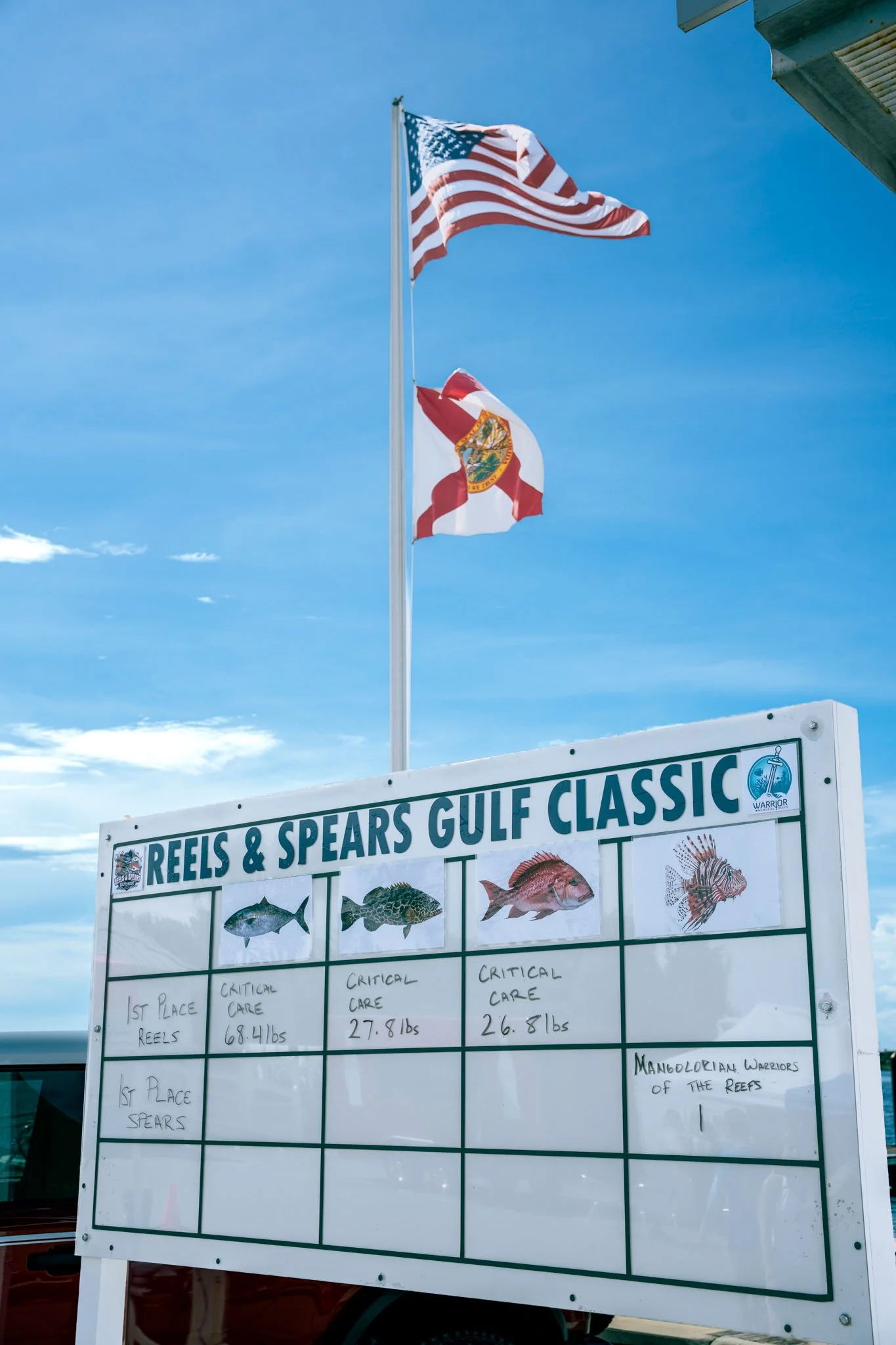 Two American flags flying on flagpoles above a sign at a fishing competition titled "Reels & Spears Gulf Classic." The sign displays images of various fish with handwritten placements and weights, judging the fish caught in the contest.