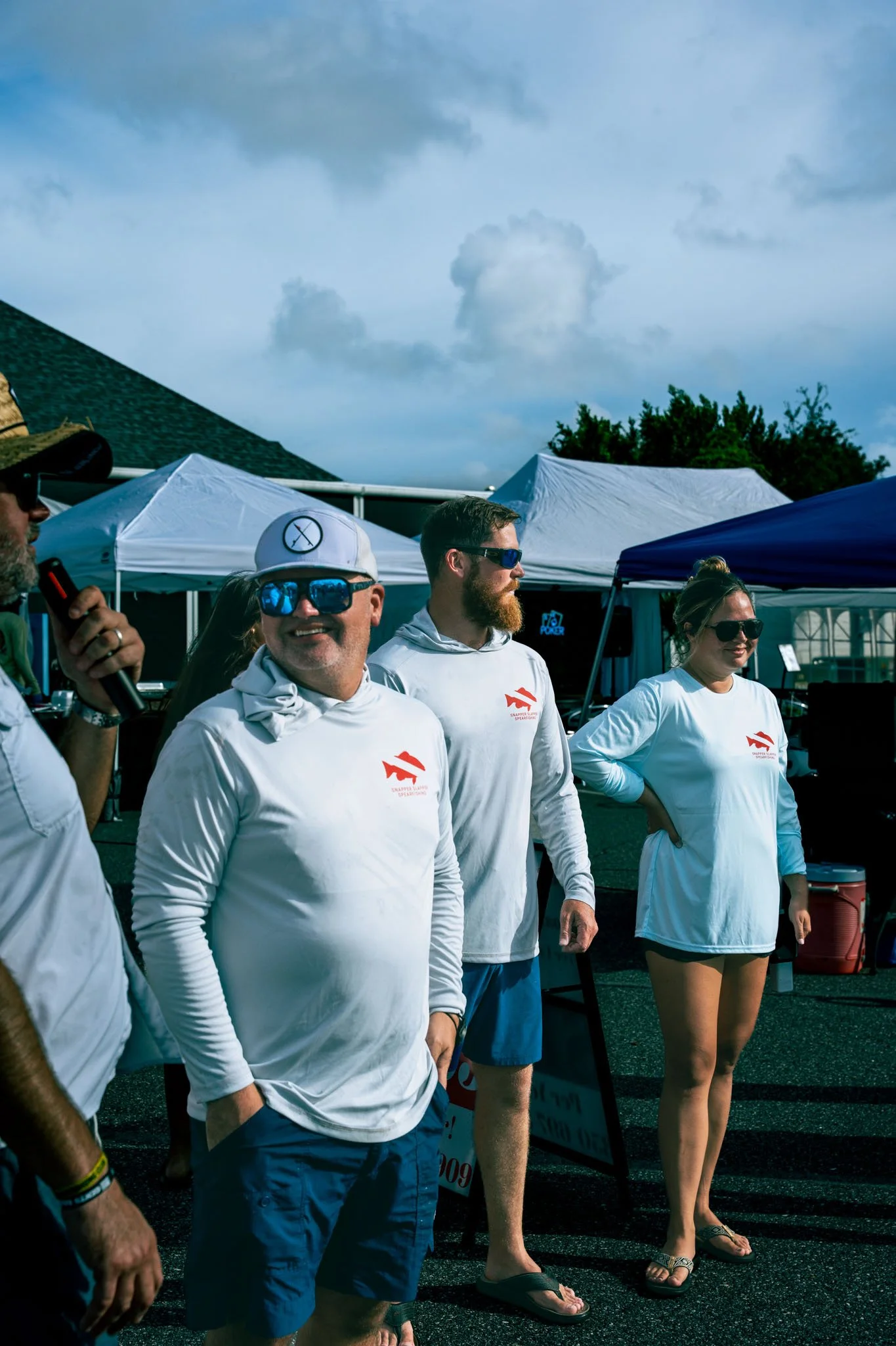 A group of people wearing white shirts with a red logo, standing outdoors among tents, with cloudy sky above.