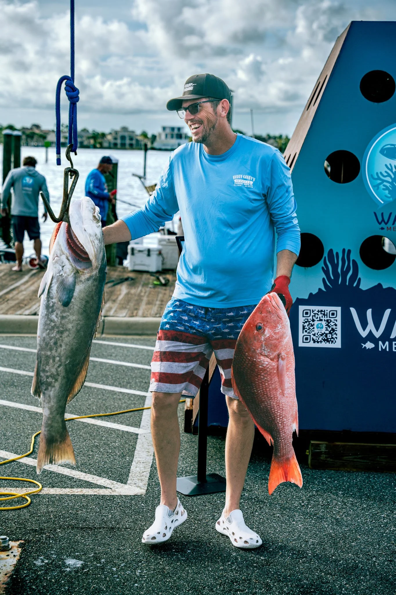 A man wearing a black cap, sunglasses, a light blue shirt, and shorts with red, white, and blue stripes, standing on a boat dock holding two large fish with a blue weatherproof sign behind him. The sign features a logo with coral and fish images and 