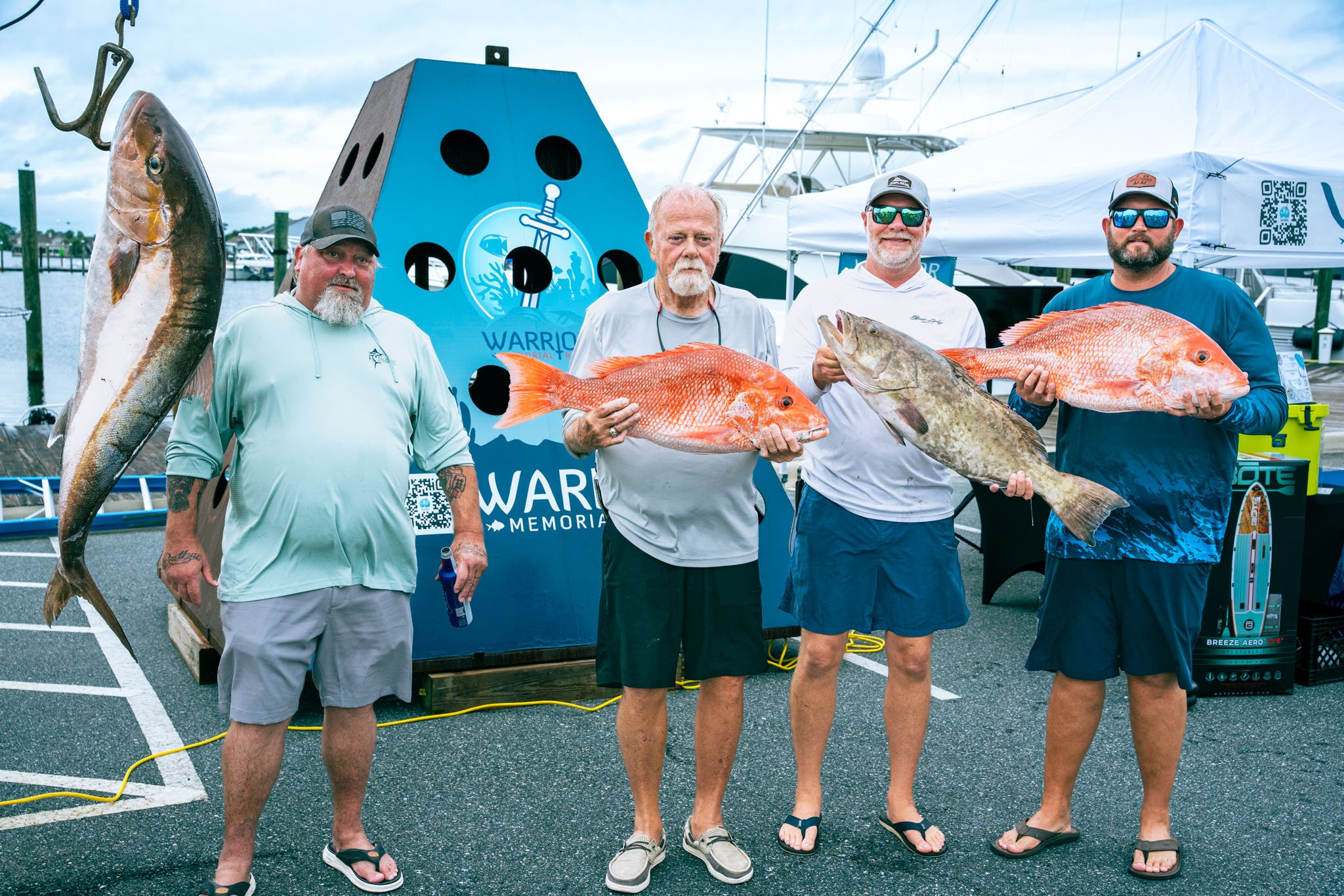 Four men standing on a dock holding large fish, with boats in the background and a blue shark silhouette sign.