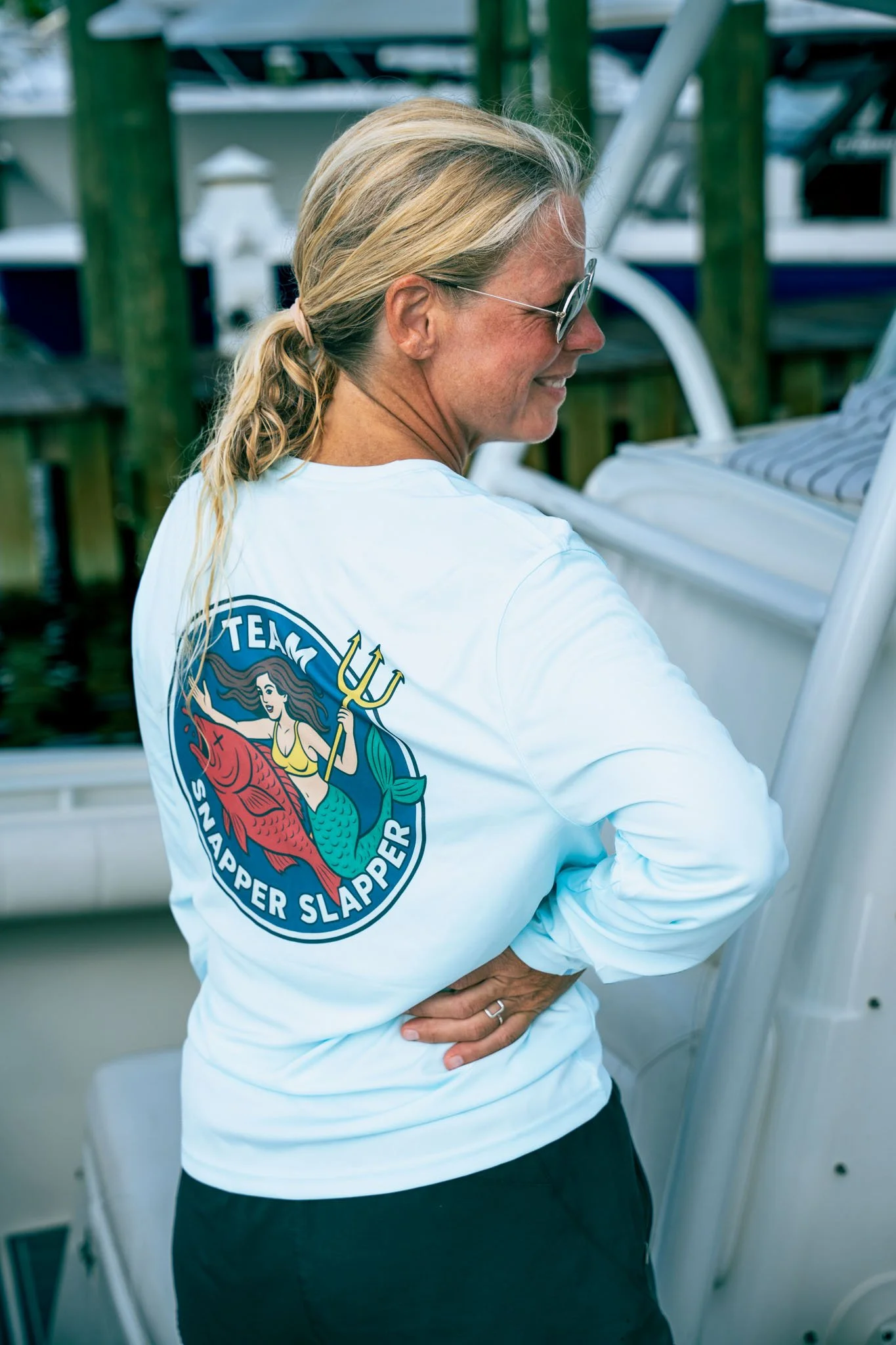A woman with blonde hair tied back, wearing sunglasses and a white shirt with a logo, stands outdoors near boats, smiling and holding her hand on her hip.