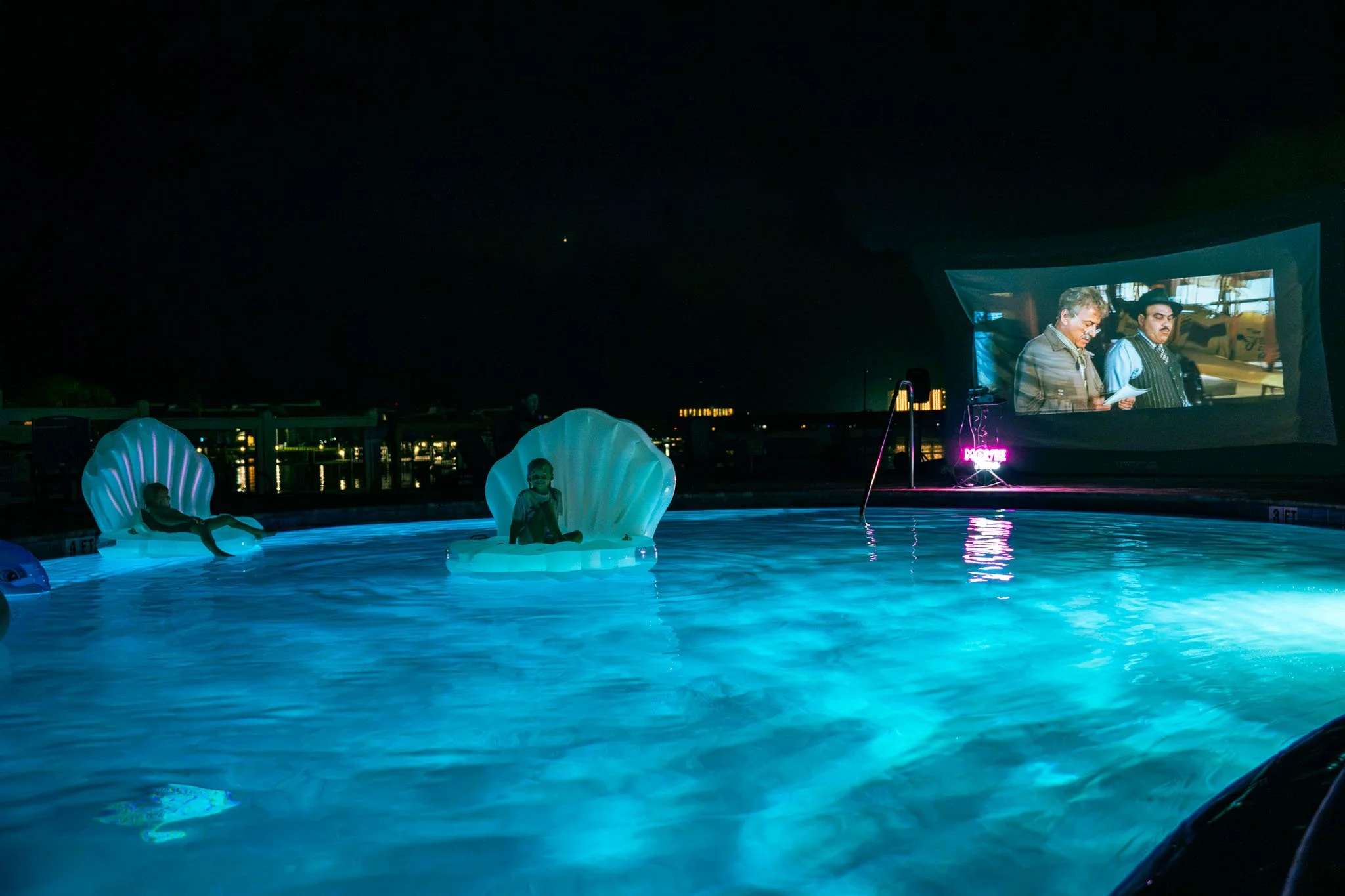 Nighttime scene at Point South Marina - Bay Point (Panama City Beach) pool with illuminated floating chairs and people sitting on them, large movie screen displaying two men reading papers, city lights in the background.