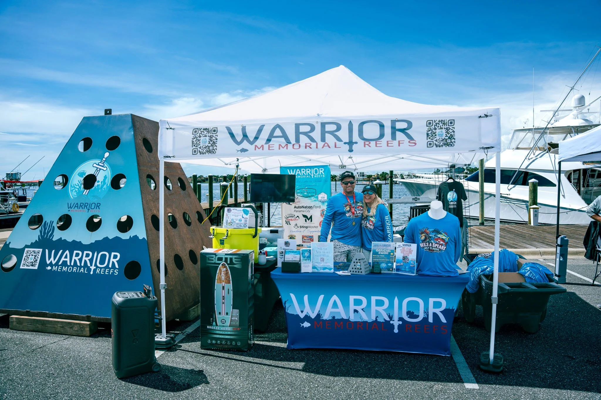 A promotional booth for Warrior Memorial Reefs at a dock with boats. The booth features a large blue and black sculpture with the company's logo, tables with merchandise including T-shirts, a TV screen, and various informational materials. Two smilin
