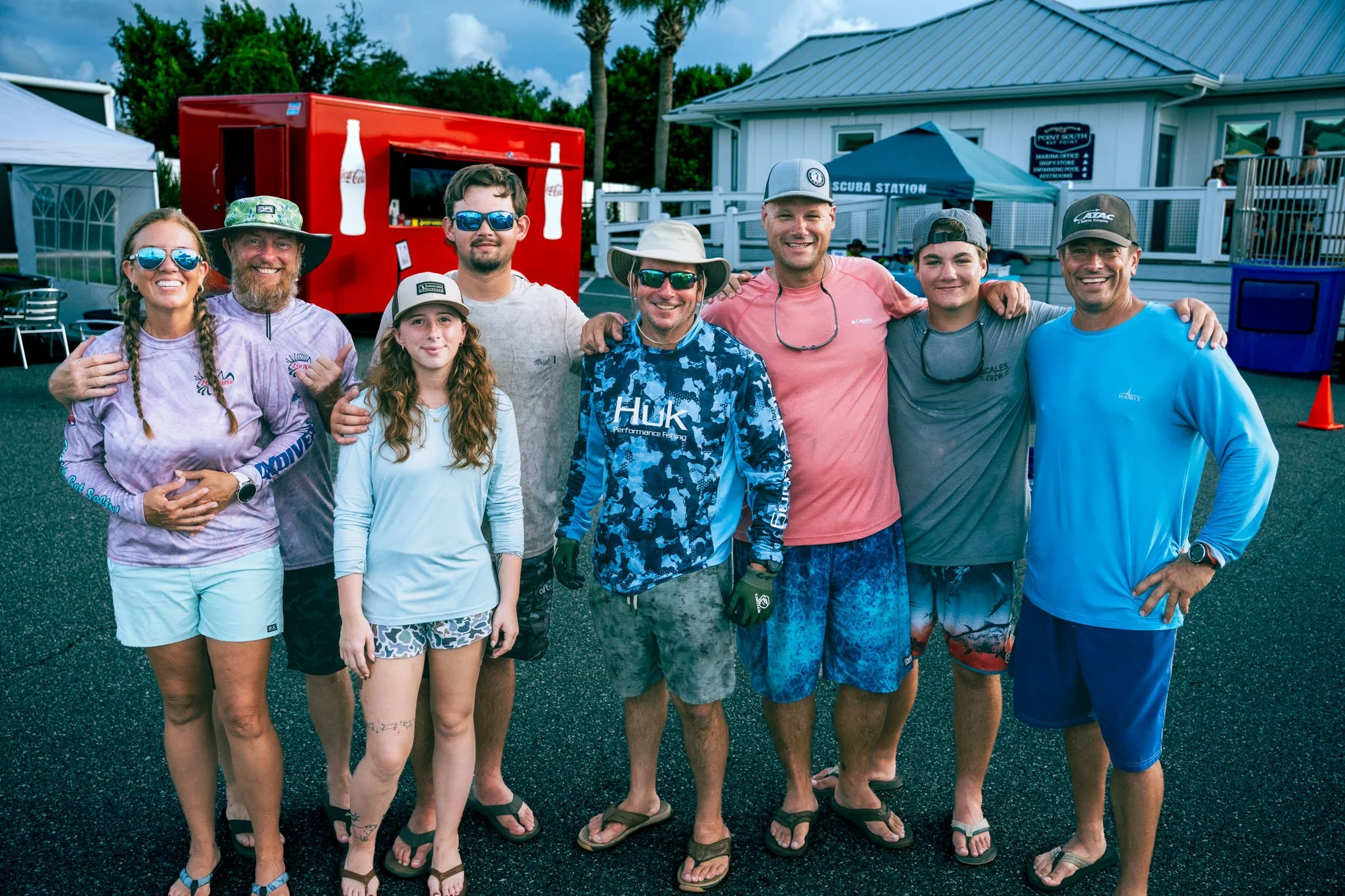 A group of nine people standing outdoors at a pool or aquatic center, smiling and posing for a photo, with pool facilities and palm trees in the background.