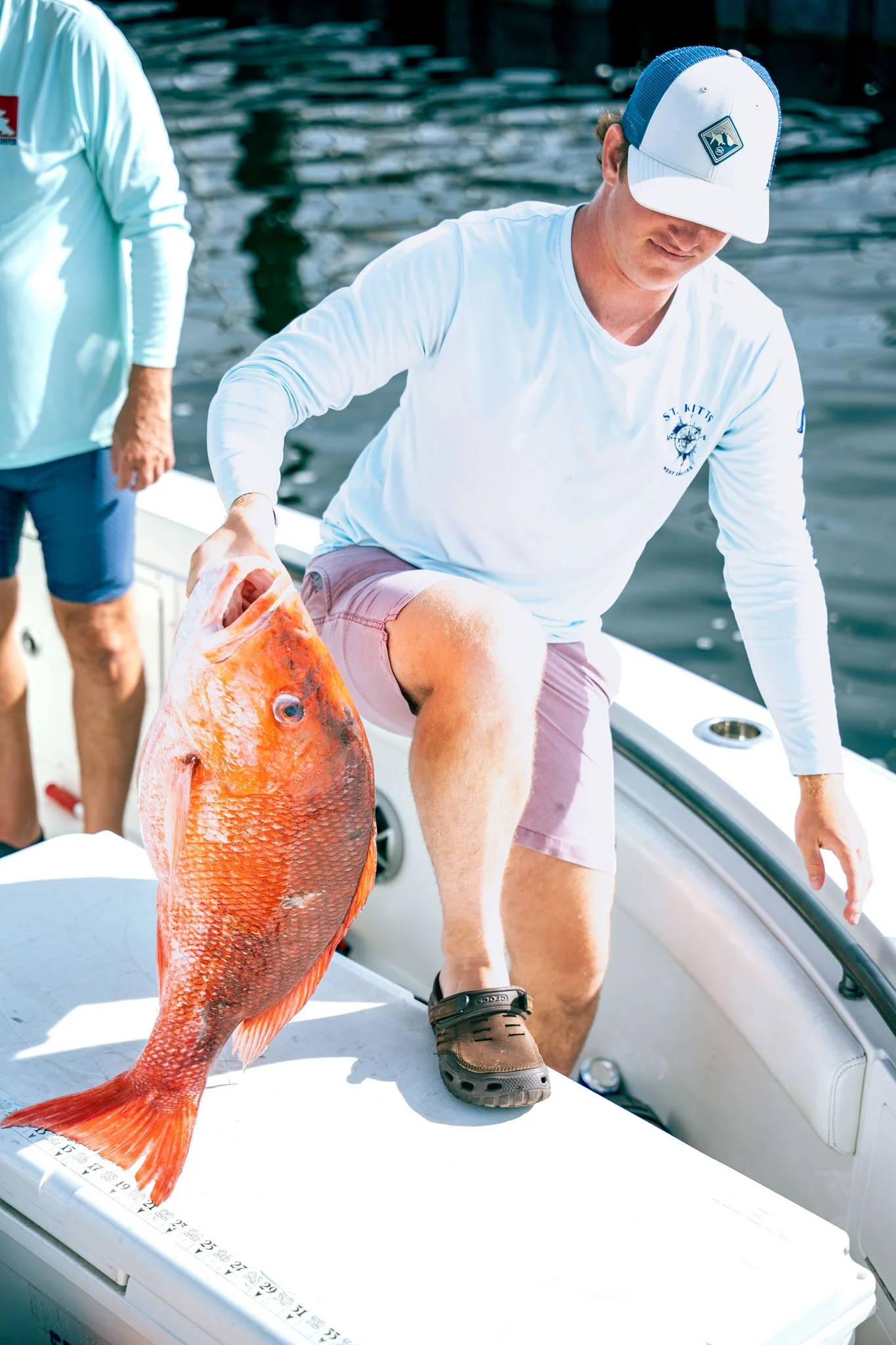 A man in a white long-sleeve shirt, pink shorts, and a blue and white cap on a boat, holding a large orange fish with red fins and scales, with another person partially visible in the background.