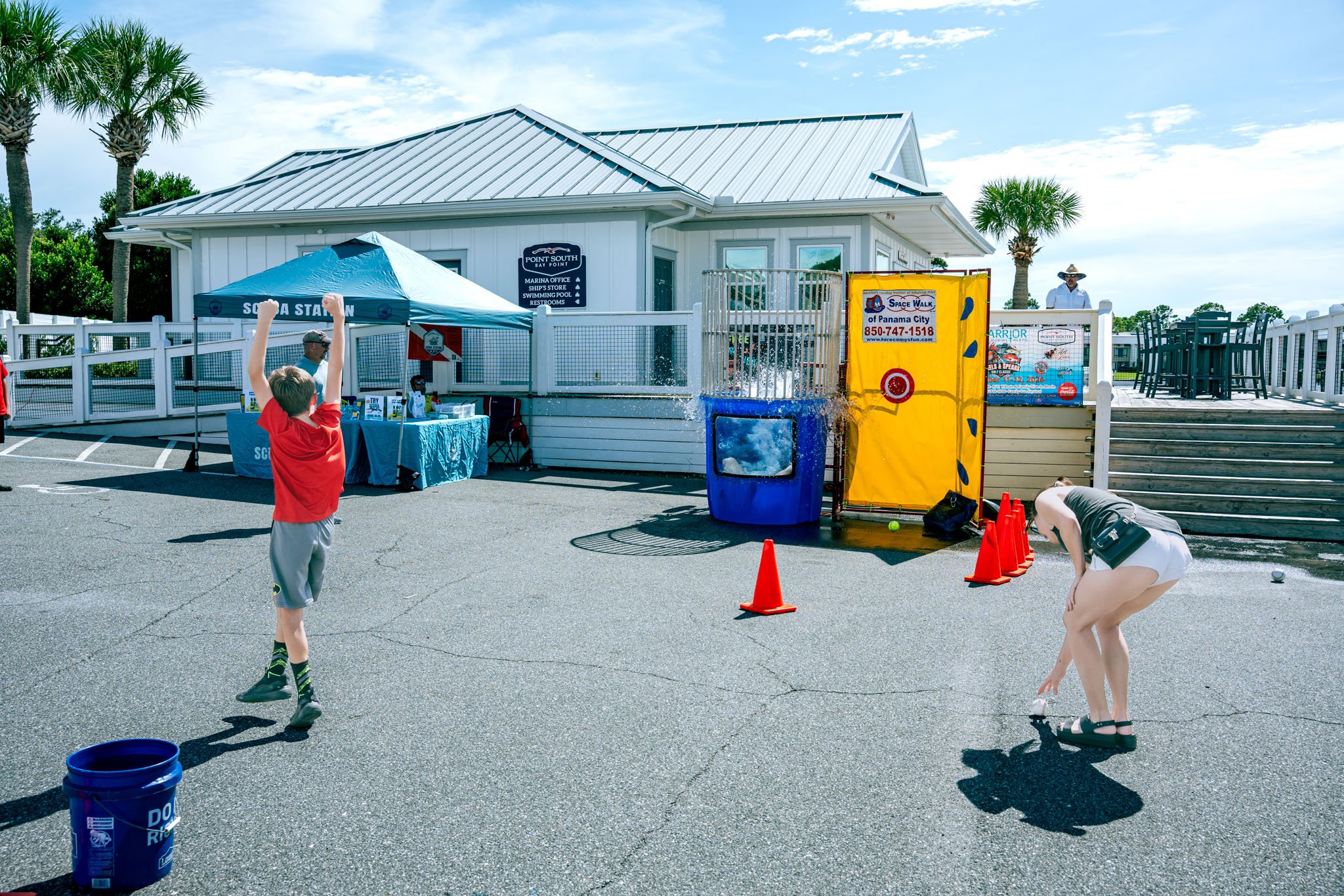 People playing shuffleboard on an outdoor court with palm trees, a white building, and a clear blue sky in the background.