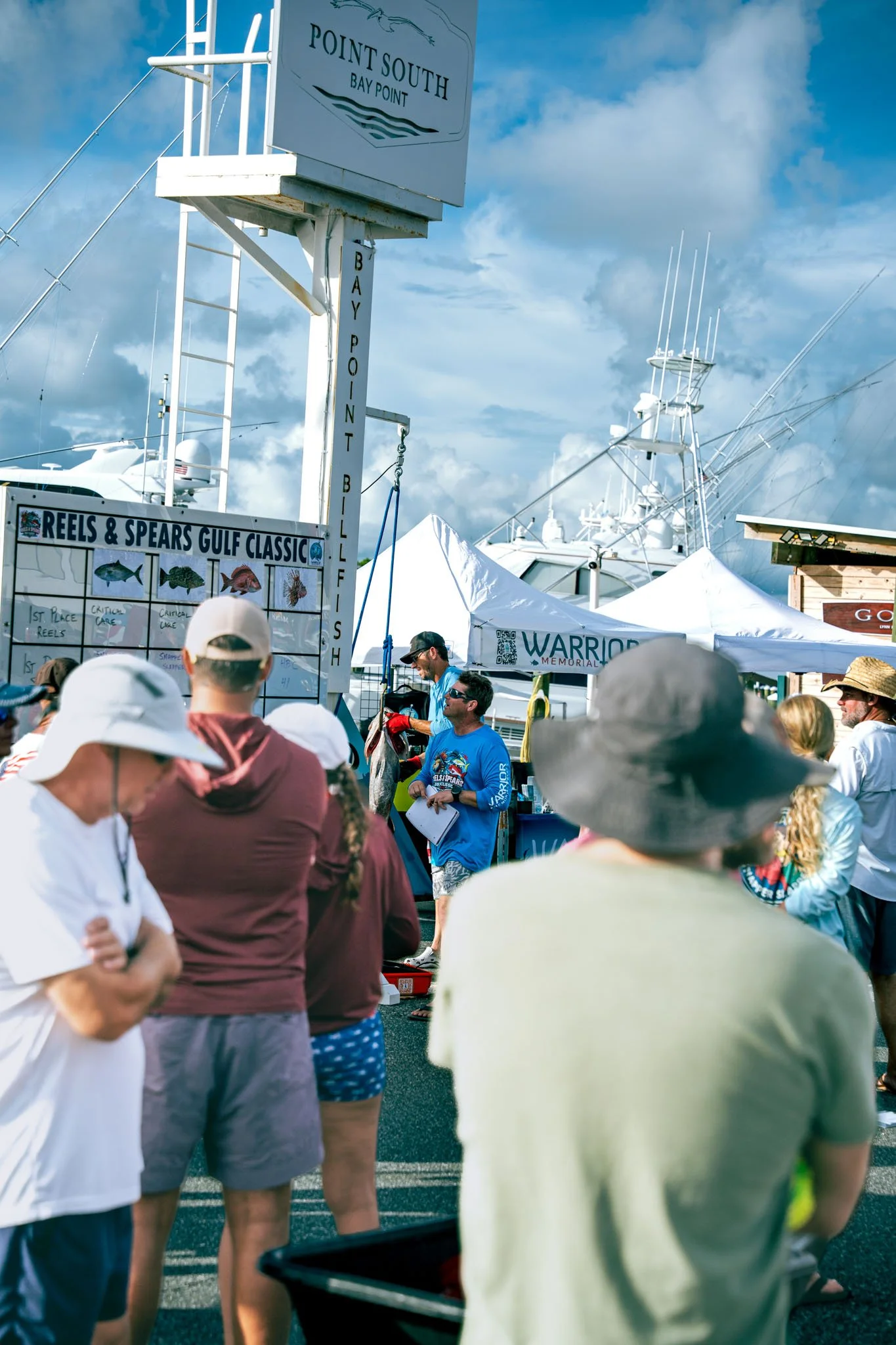 Crowd of people at the Warrior Memorial Reefs Reels and Spears Fishing Tournament event at Point South Marina - Bay Point (Panama City Beach) with white tents, a sign for Point South Bay Point, and boats in the background under a partly cloudy sky.