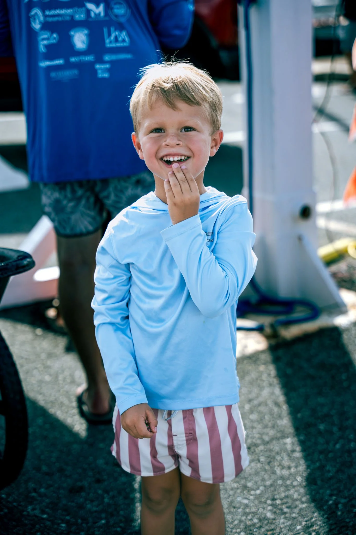 A young boy with blond hair smiling and touching his chin, wearing a light blue long-sleeve shirt and red and white striped shorts, standing outdoors near a motorcycle with a blurred background of people and structures.