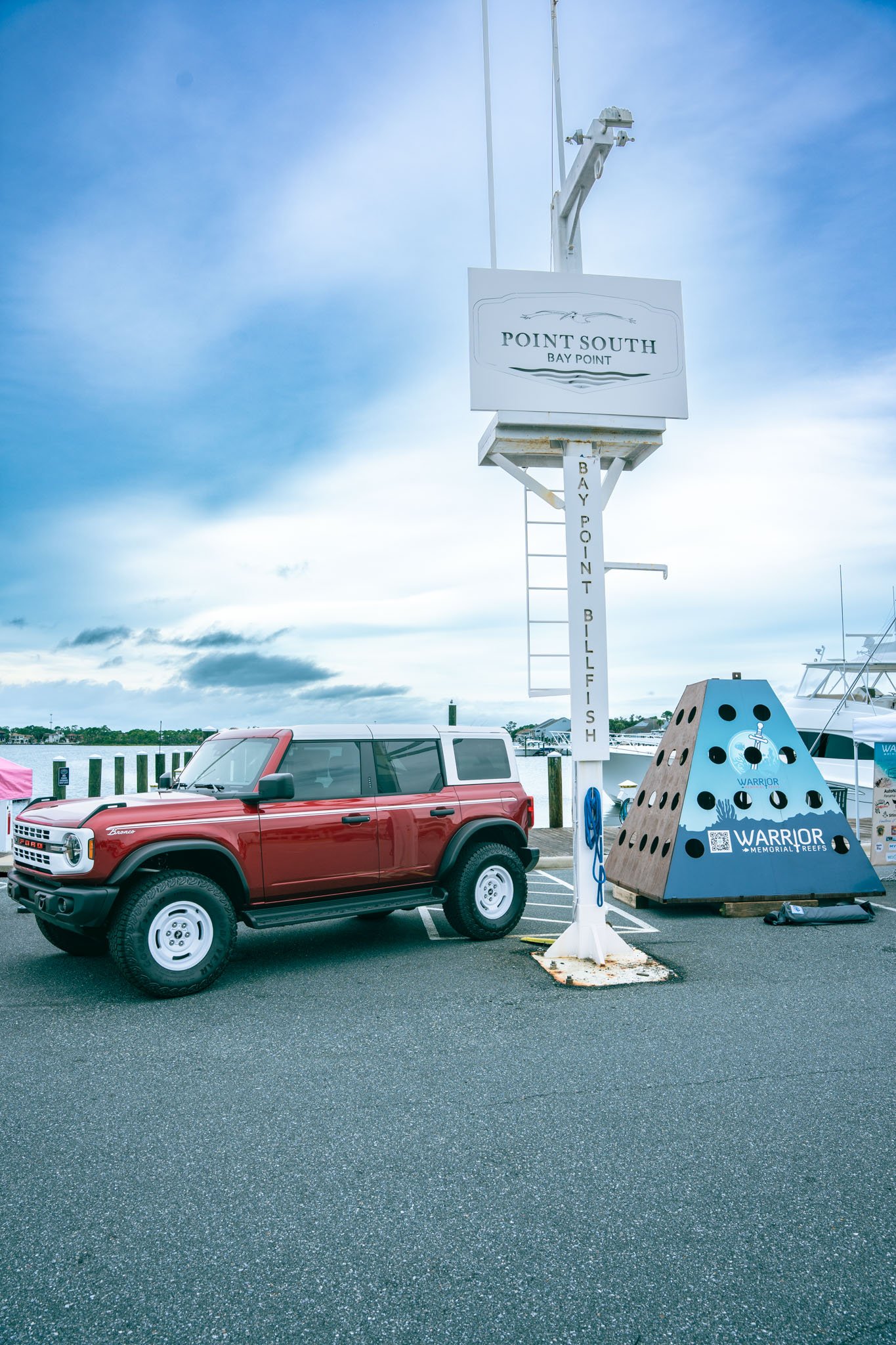 A red Ford Bronco parked in a marina parking lot, with a sign reading 'Point South Bay Point' and a memorial art piece with the word 'Warrior' beside it. The sky is partly cloudy.