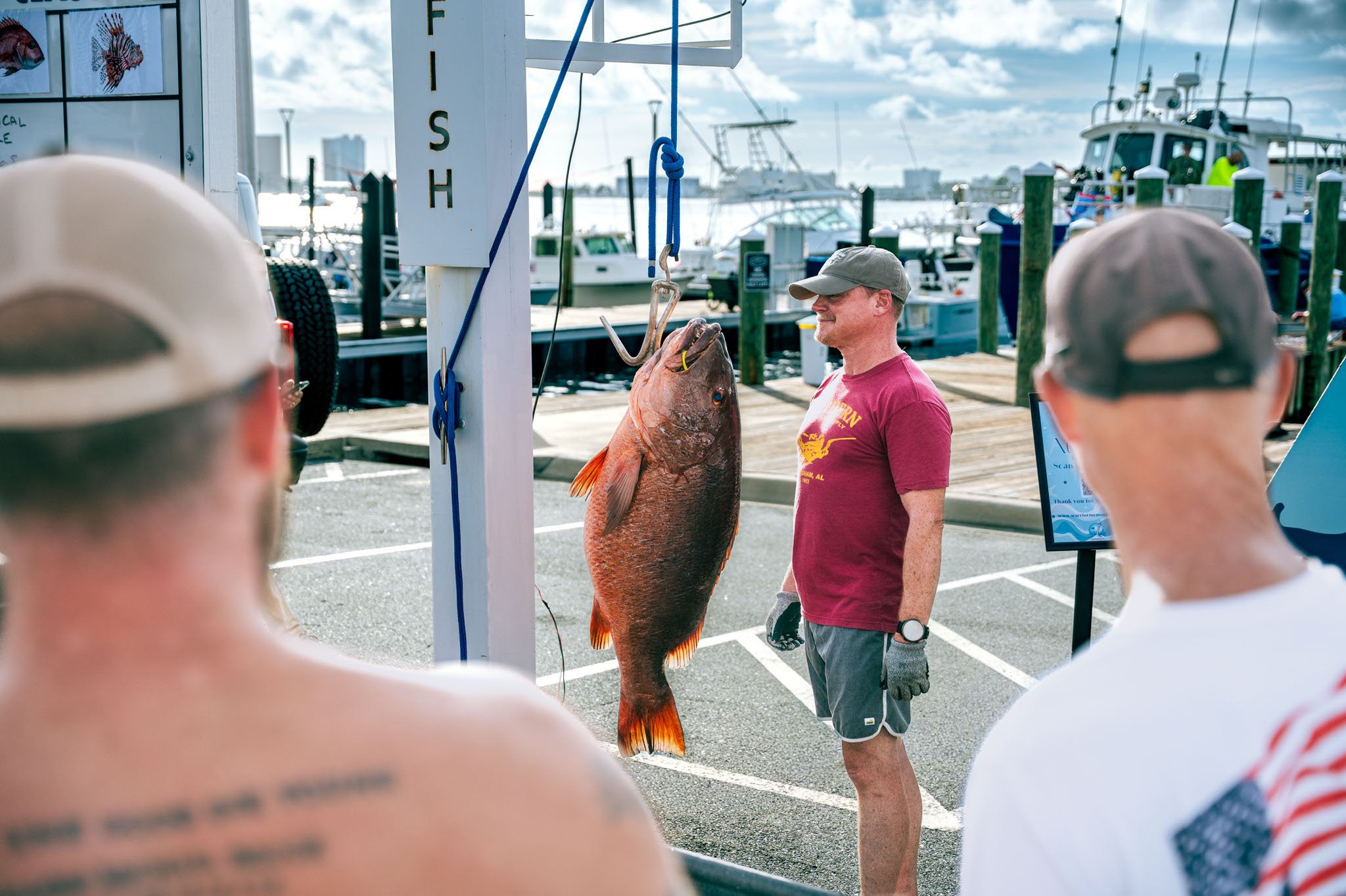 A man with a beard and gray cap stands next to a large cubera snapper hanging from a scale at a marina. Several people are in the foreground, with boats docked in the background under a partly cloudy sky.