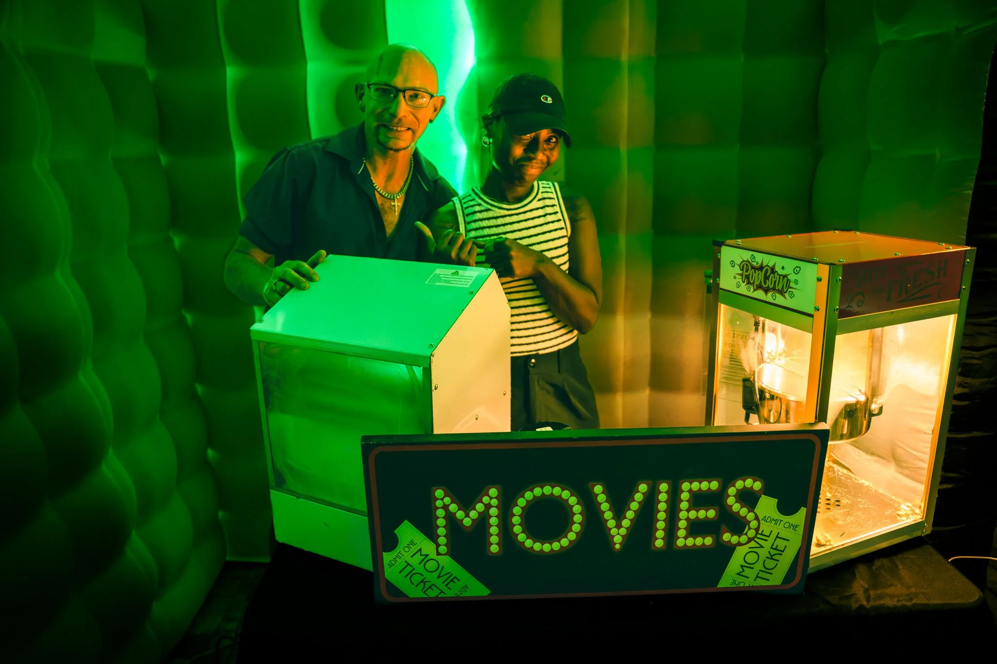 Two people posing in a green-lit setting, standing near a popcorn machine and movie ticket sign.