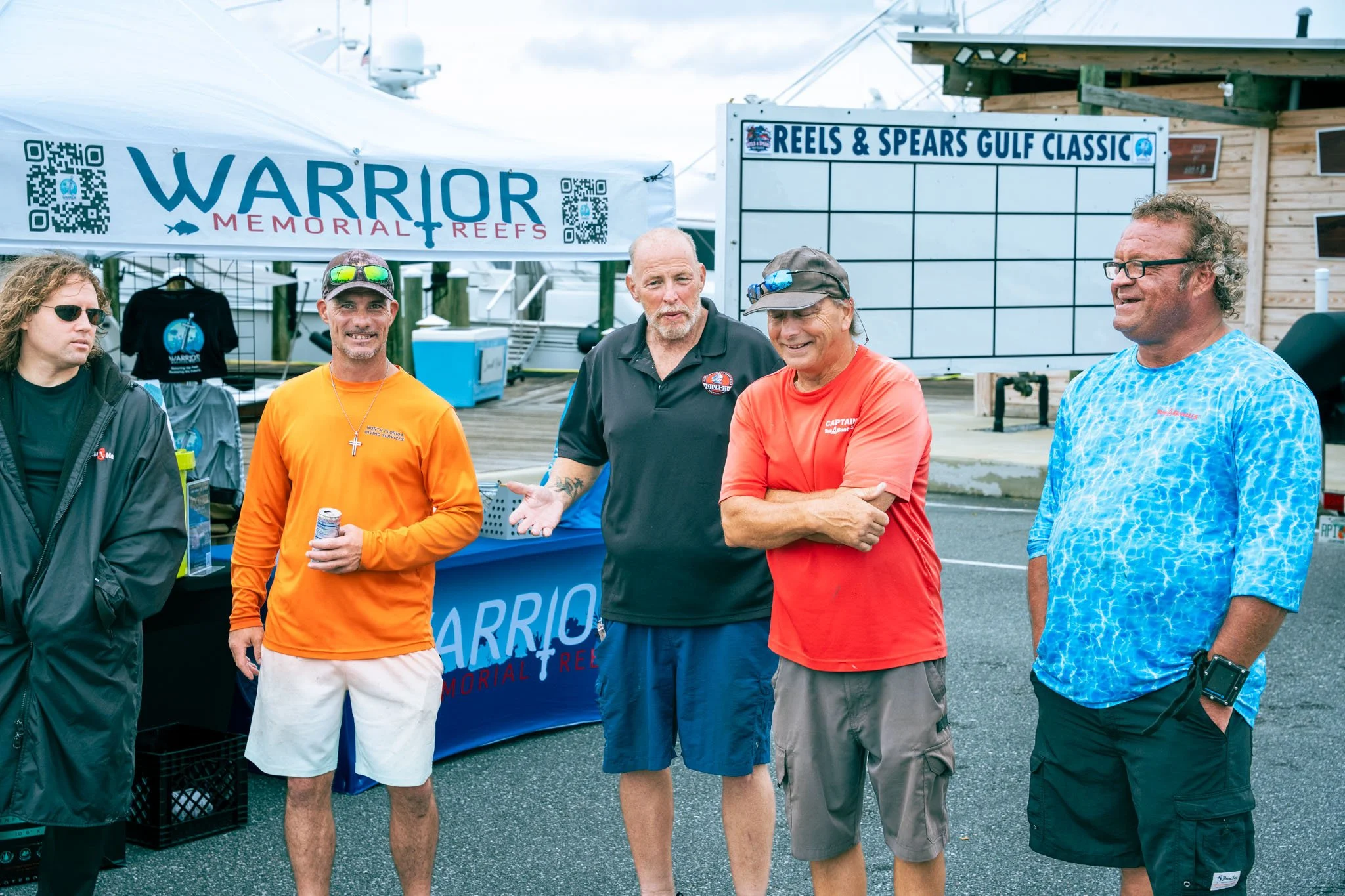 Group of five men standing in front of a tent and a sign that reads 'Reels & Spears Gulf Classic'. The men are wearing casual shirts and shorts, some with sunglasses, and are smiling or talking. The background shows a wooden building and cloudy sky.